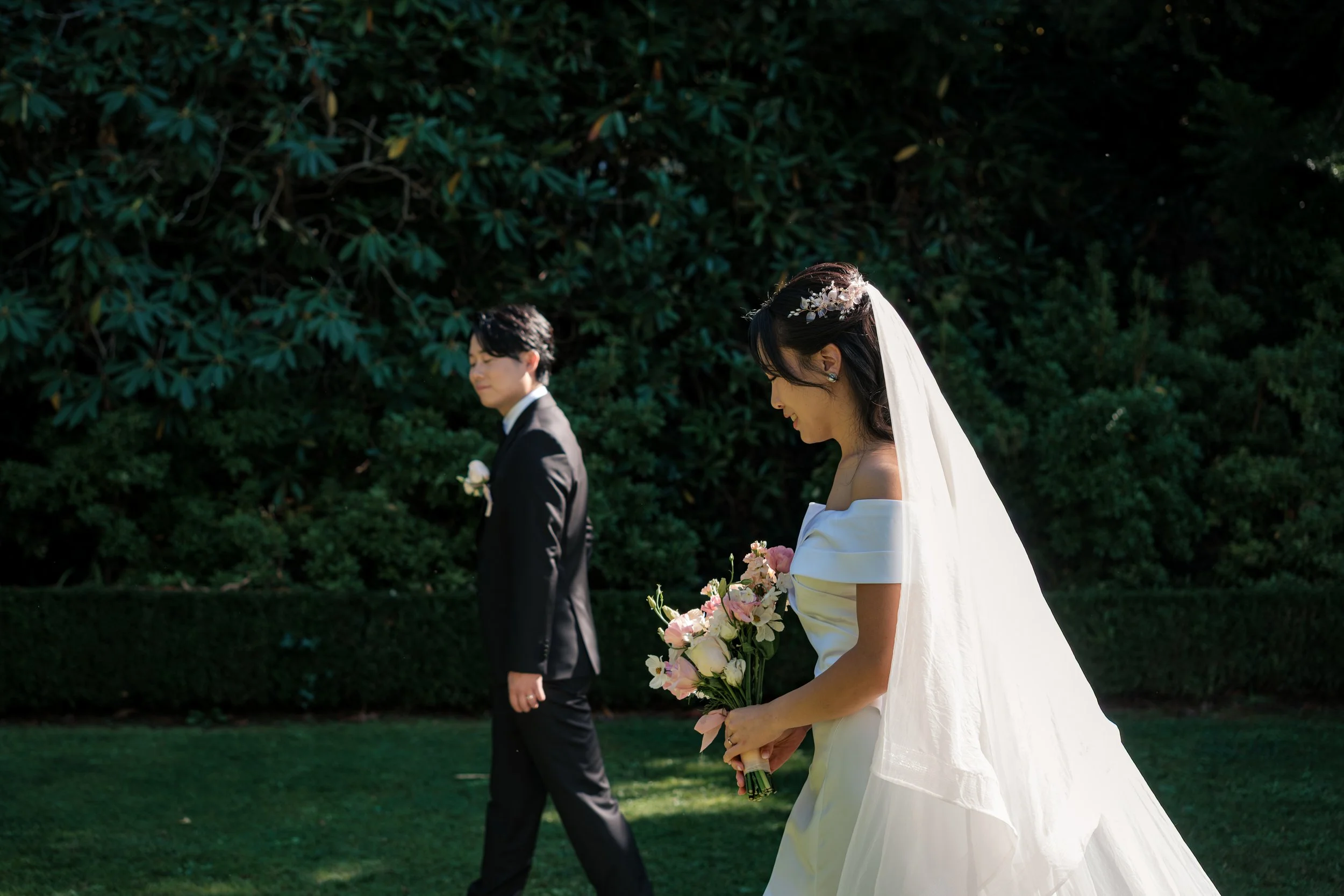 Bride in a white wedding dress holding a bouquet of pink and white flowers, walking outdoors with the groom in a black suit walking behind her in a garden setting.