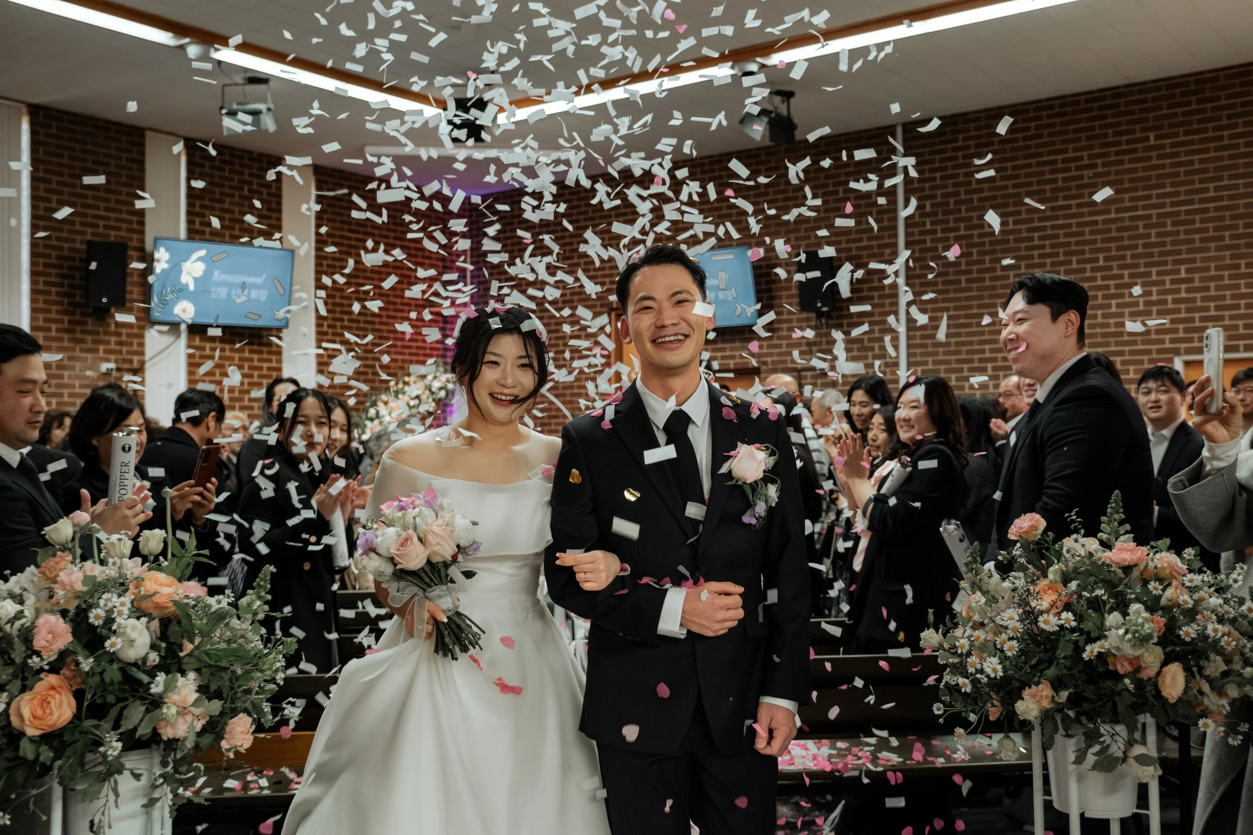 A newly married couple walking down the aisle after their wedding ceremony surrounded by friends and family, with confetti falling around them.