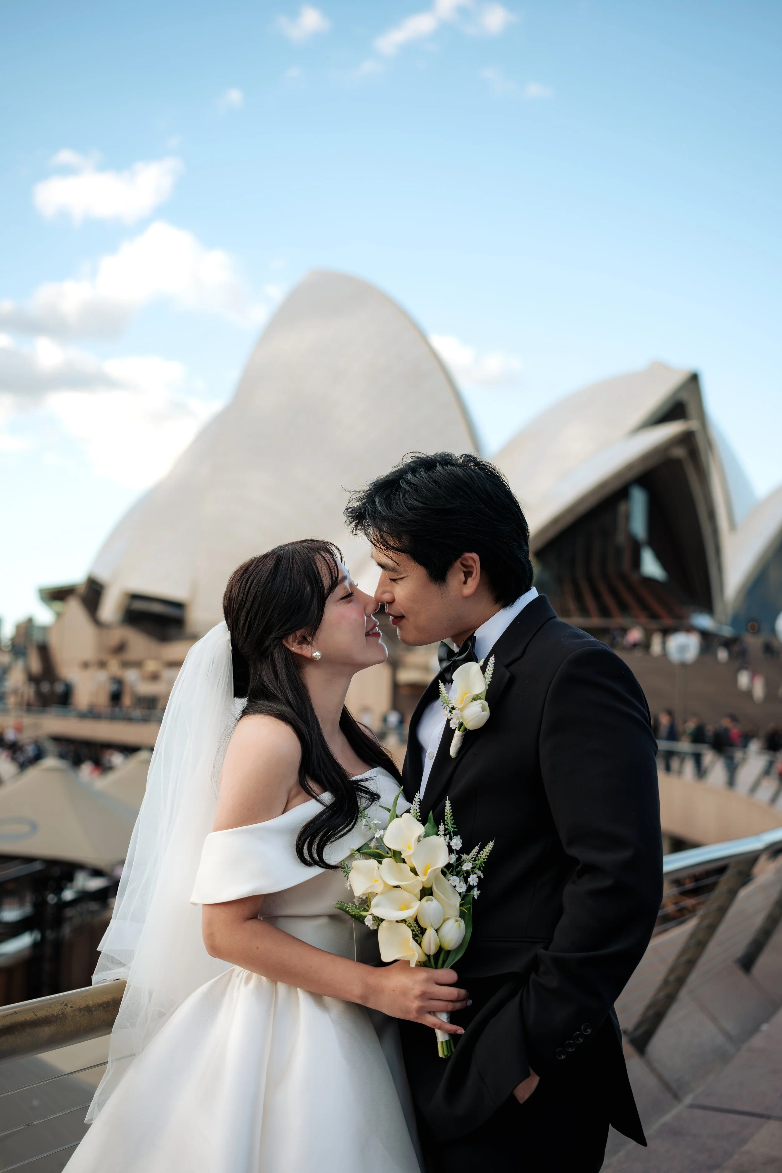 A bride and groom are close together, touching noses, with the Sydney Opera House in the background.