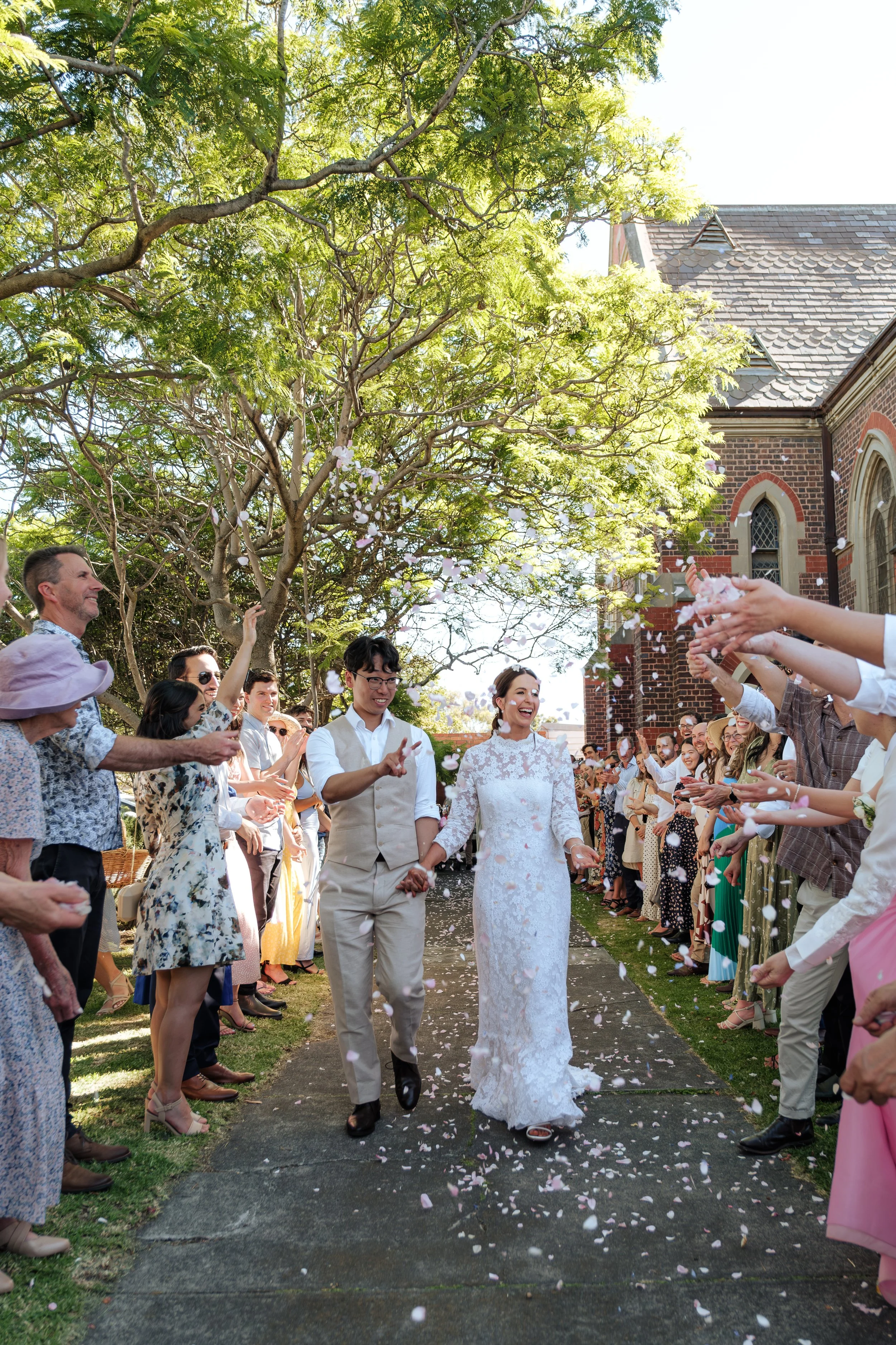 A newlywed couple walking down the aisle, surrounded by guests throwing flower petals during daytime outdoor wedding ceremony at a church.