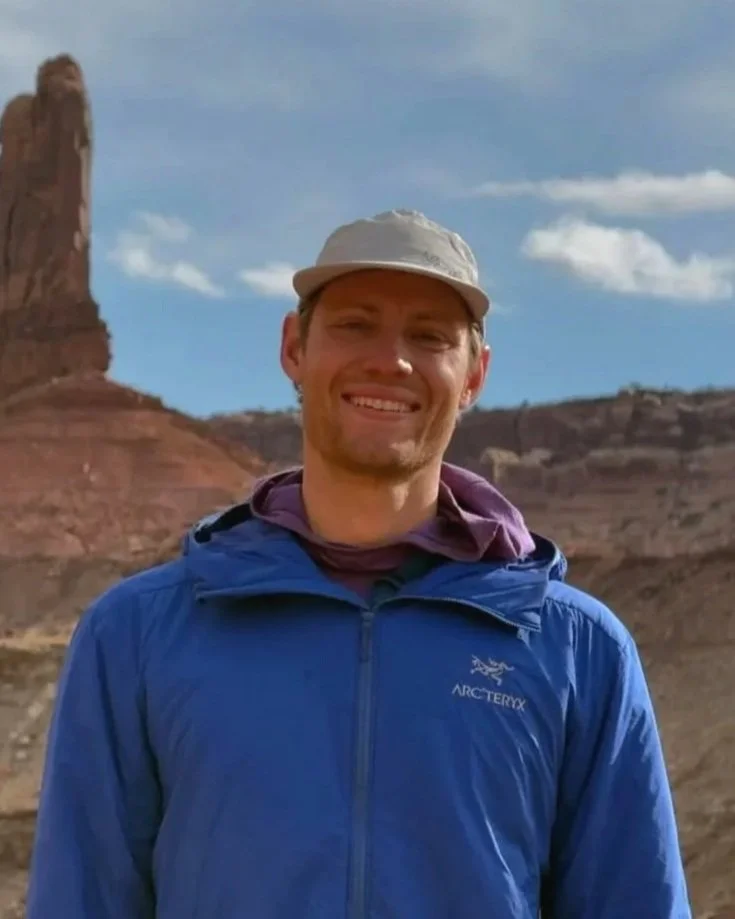 A smiling man wearing a beige cap and a blue Arc'teryx jacket stands outdoors with a desert landscape and rocks behind him.