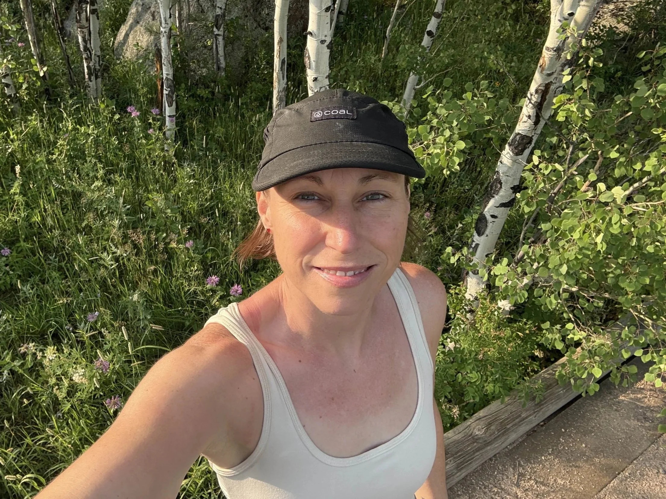 A woman with fair skin, brown hair, and blue eyes smiling at the camera in a white tank top, wearing a black cap with the COAL logo, outdoors among green trees and grass.