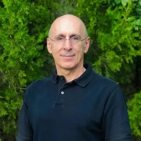 A middle-aged man with glasses and a bald head, wearing a dark collared shirt, standing outdoors in front of green foliage.