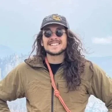 Man with long hair, wearing sunglasses, a cap, and a brown jacket outdoors, smiling at the camera.