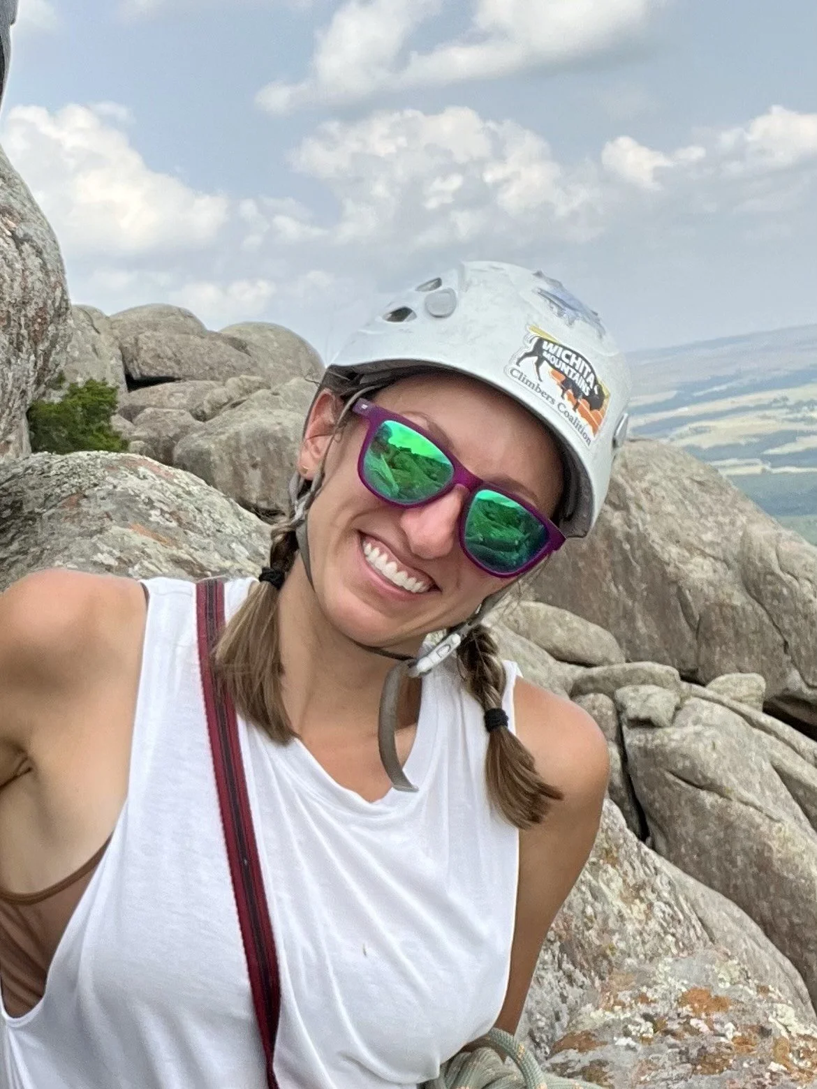 A woman wearing a helmet and sunglasses, smiling while climbing rocks on a mountain summit.