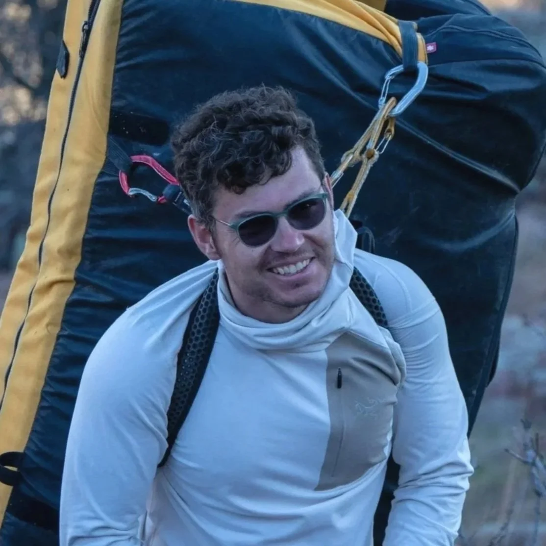 Man outdoors wearing sunglasses, a white jacket, and a backpack, carrying a large yellow and black gear or bag on his back.