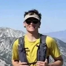 Person smiling outdoors with mountains and clear sky in background, wearing sunglasses, a yellow shirt, and a backpack.