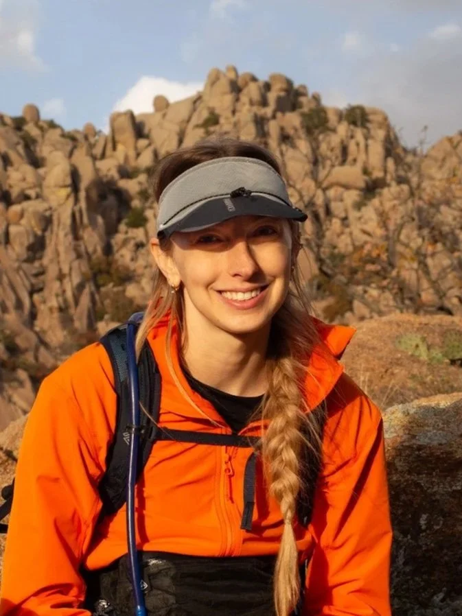 Young woman outdoors in hiking gear, smiling, with a rocky mountain landscape in the background.