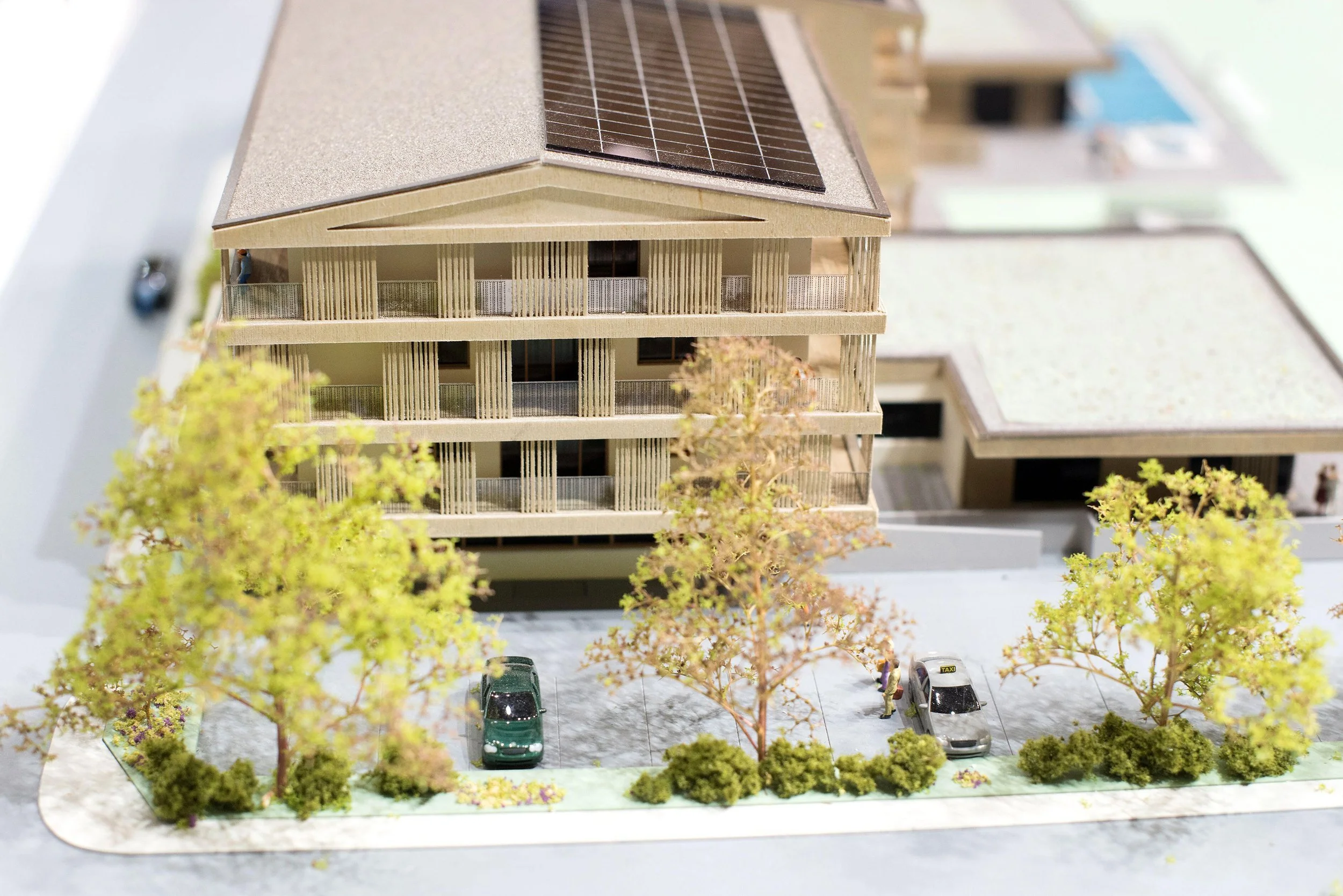 A detailed architectural model of a multi-story residential building with a sloped roof that has solar panels. The building features multiple balconies and is surrounded by miniature trees and parked cars.
