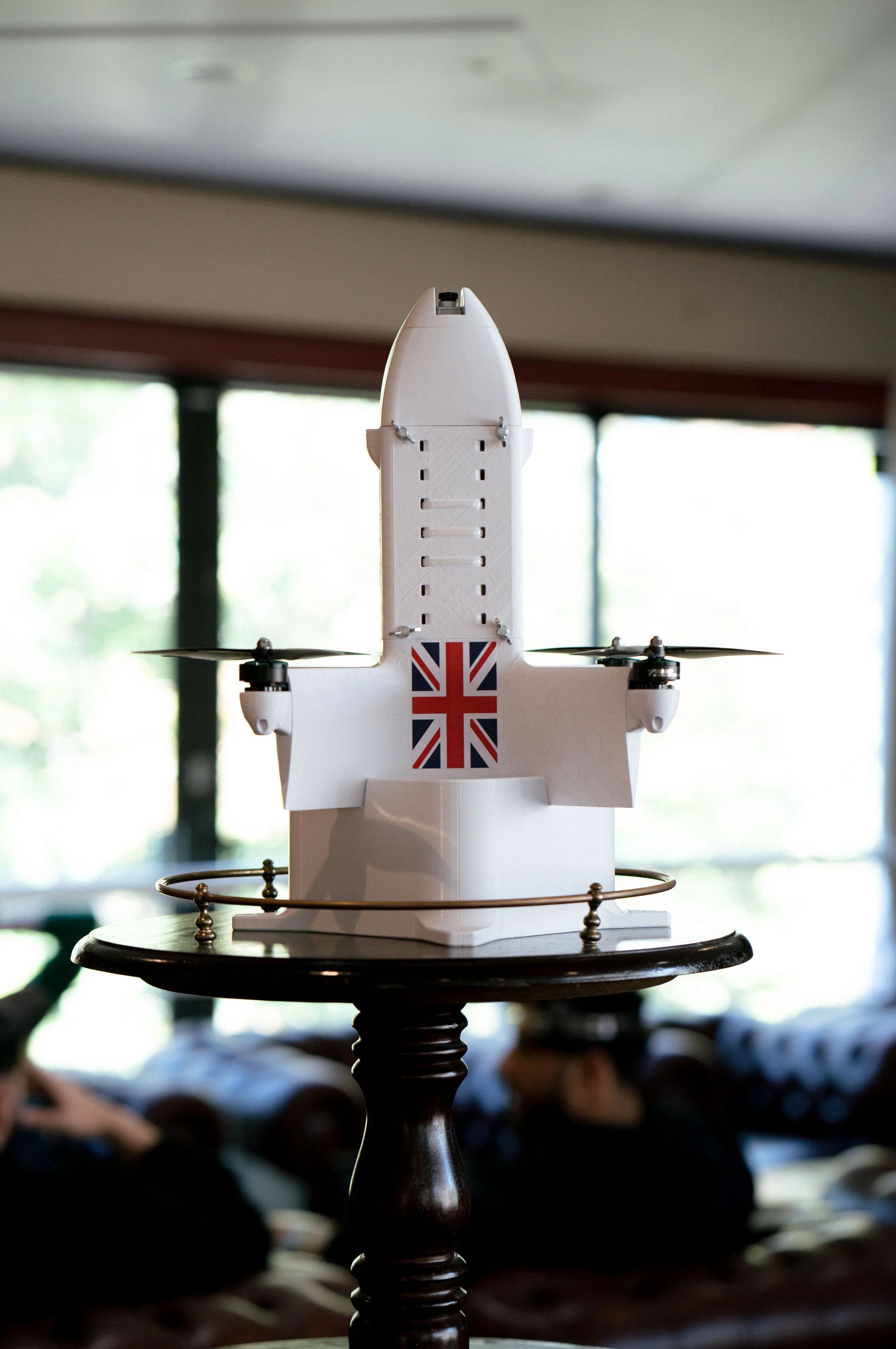 A white drone with a UK flag decal on its underside, placed on a wooden table with a metal railing around it, indoor setting with blurred background of people and windows.
