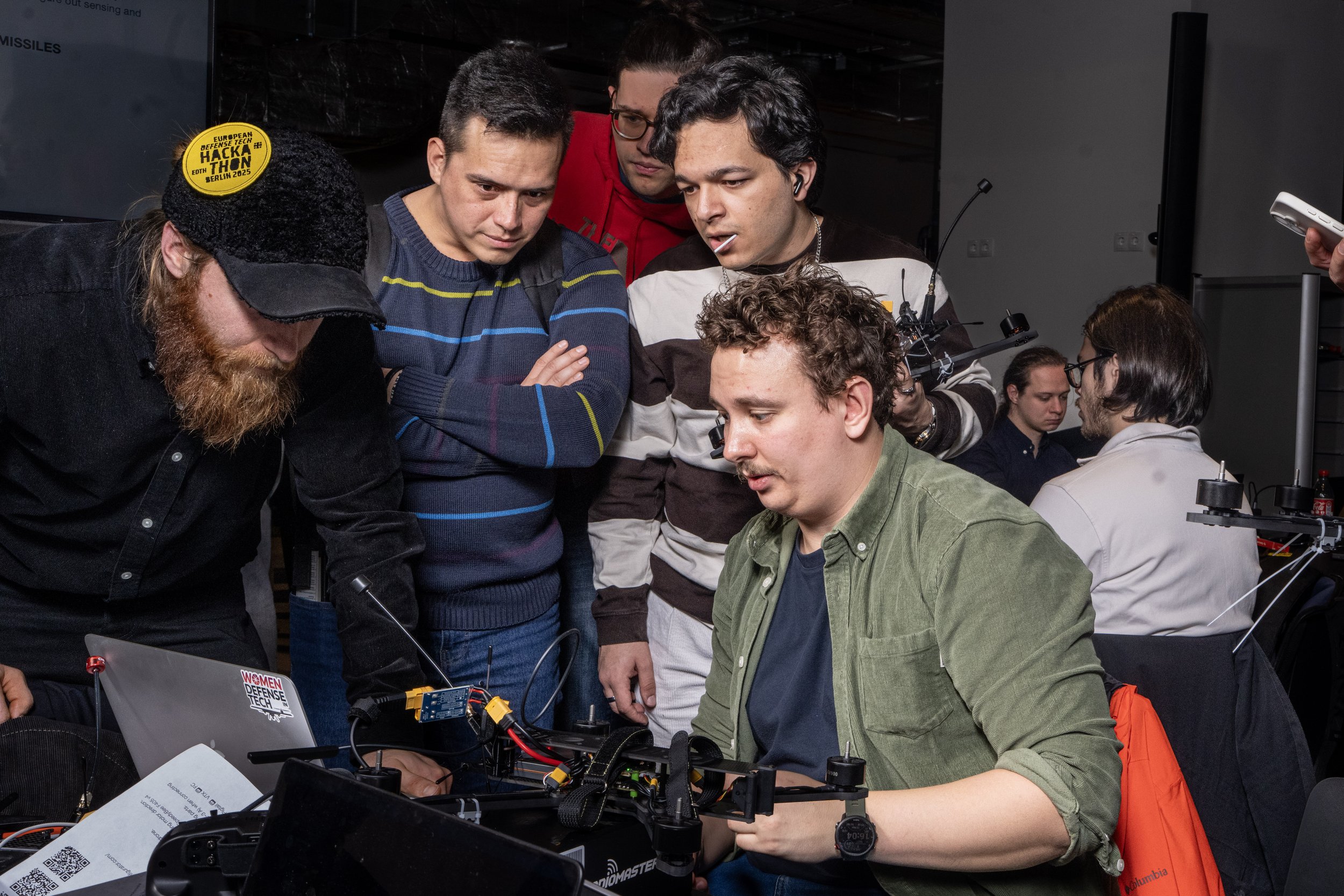 A group of young adults gathered around a table working on a robot or electronic device, with one person seated and the others standing closely observing.