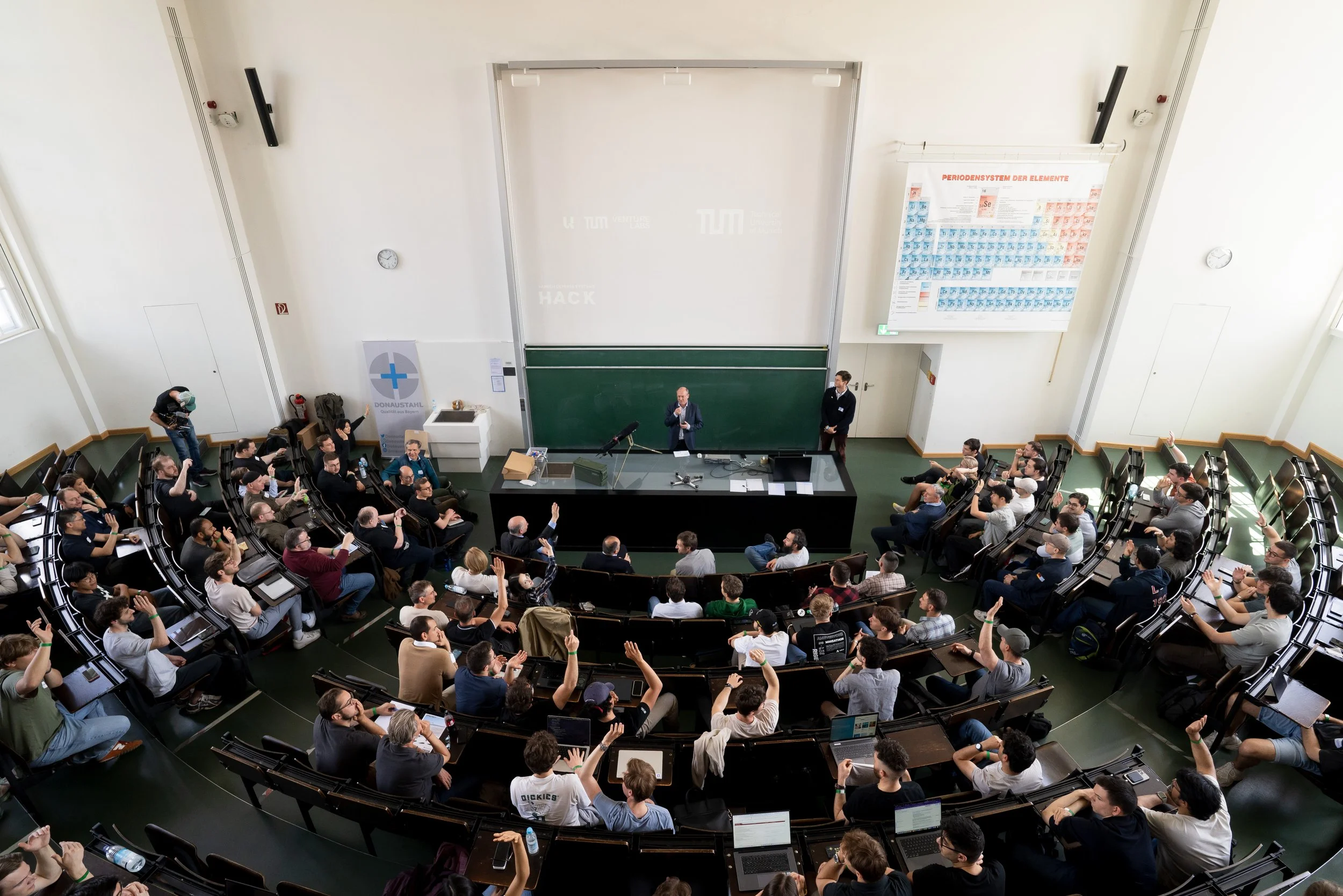 A large lecture hall filled with students and attendees, some raising their hands, listening to a speaker at the front lectern near a chalkboard, with a large periodic table poster on the wall.