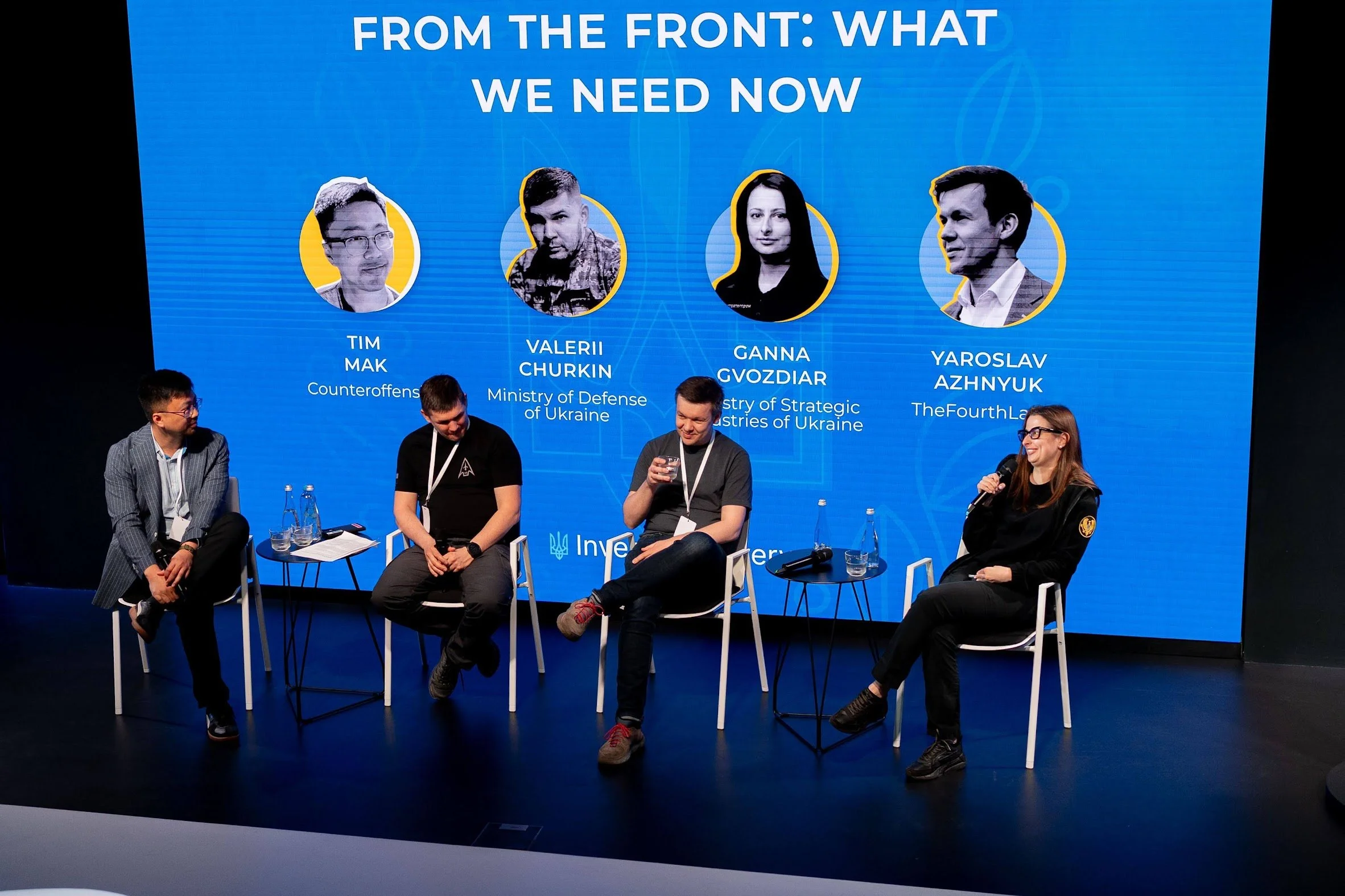 Panel discussion at a conference with four panelists sitting on stage and a large screen behind them displaying the titles and images of four speakers. The screen reads: 'From the front: What we need now,' with headshots and names of Tim Mak, Valerii Churkin, Ganna Gvozdiar, and Yaroslav Azhnyuk, representing organizations related to Ukraine.