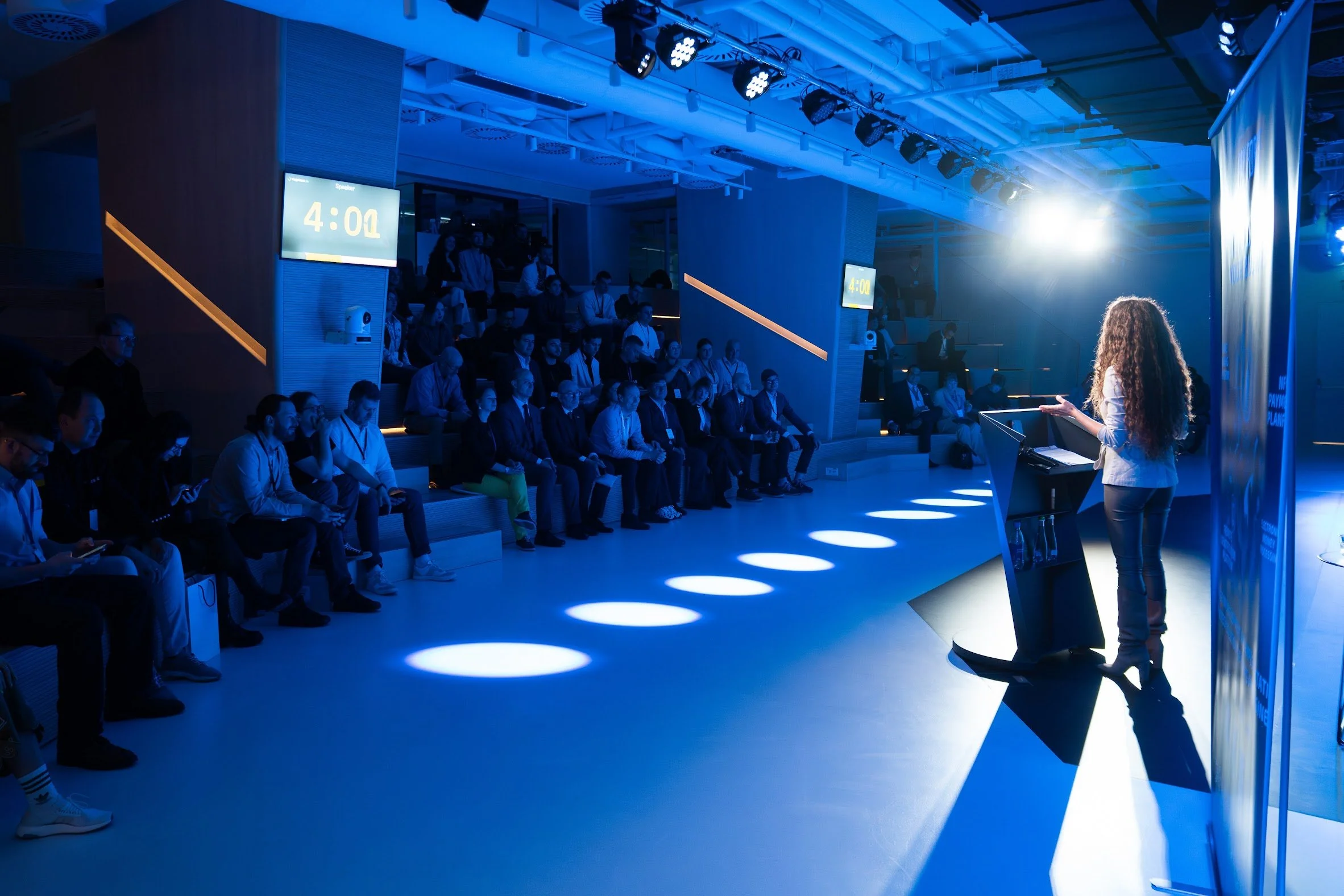 A woman giving a presentation to an audience in a dimly lit auditorium with blue lighting. The audience is seated in tiered rows, watching her at a lectern. Two digital clocks display 4:00 and 4:02.