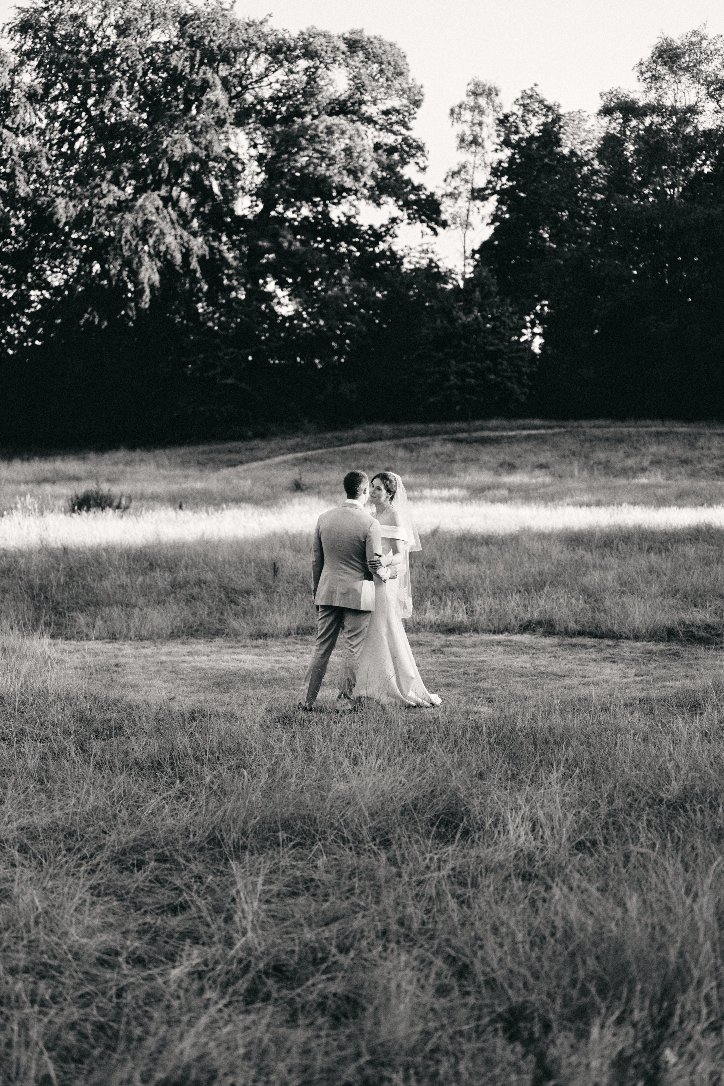 A black and white photograph of a groom and bride standing closely in a grassy field, surrounded by trees, with the bride wearing a veil and off-the-shoulder dress and the groom in a suit.