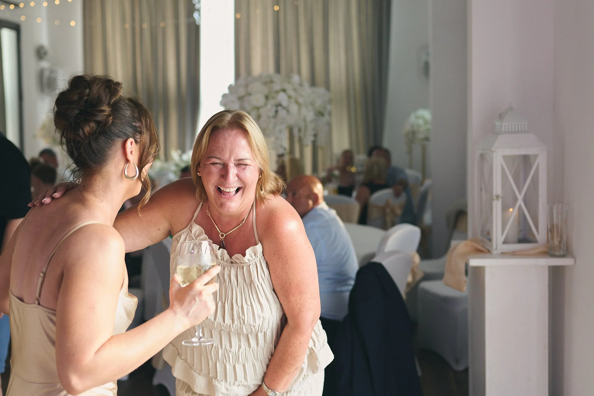 Two women sharing a joyful moment at a celebration, one holding a glass of white wine.