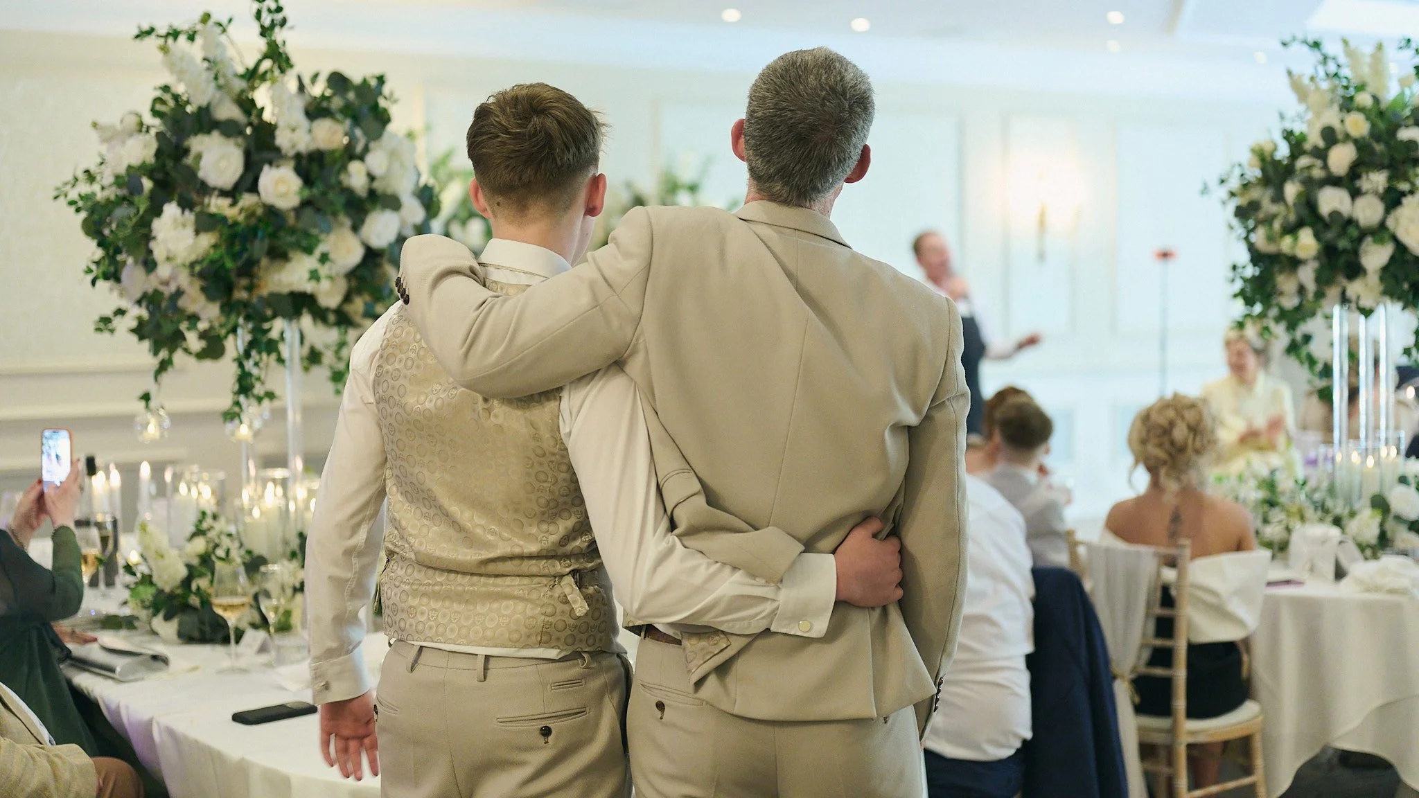 Two men standing close together, one with his arm around the other at a wedding reception, surrounded by guests and floral decorations.