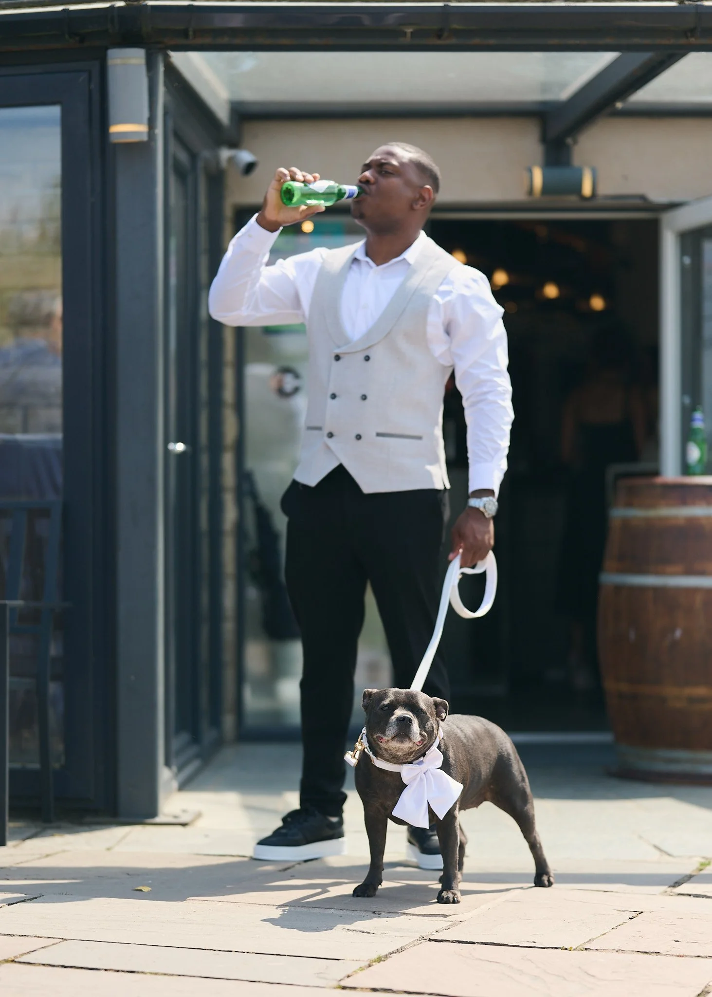 A man in a white shirt and vest is standing outside, drinking from a green bottle. A black and white dog with a white scarf around its neck is standing nearby on a leash, looking at the camera.