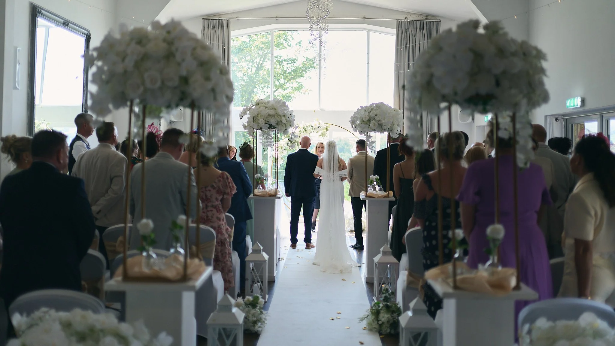 A wedding ceremony with the bride and groom standing at the altar surrounded by seated guests, decorated with large white floral arrangements and lanterns along the aisle, inside a bright indoor venue.