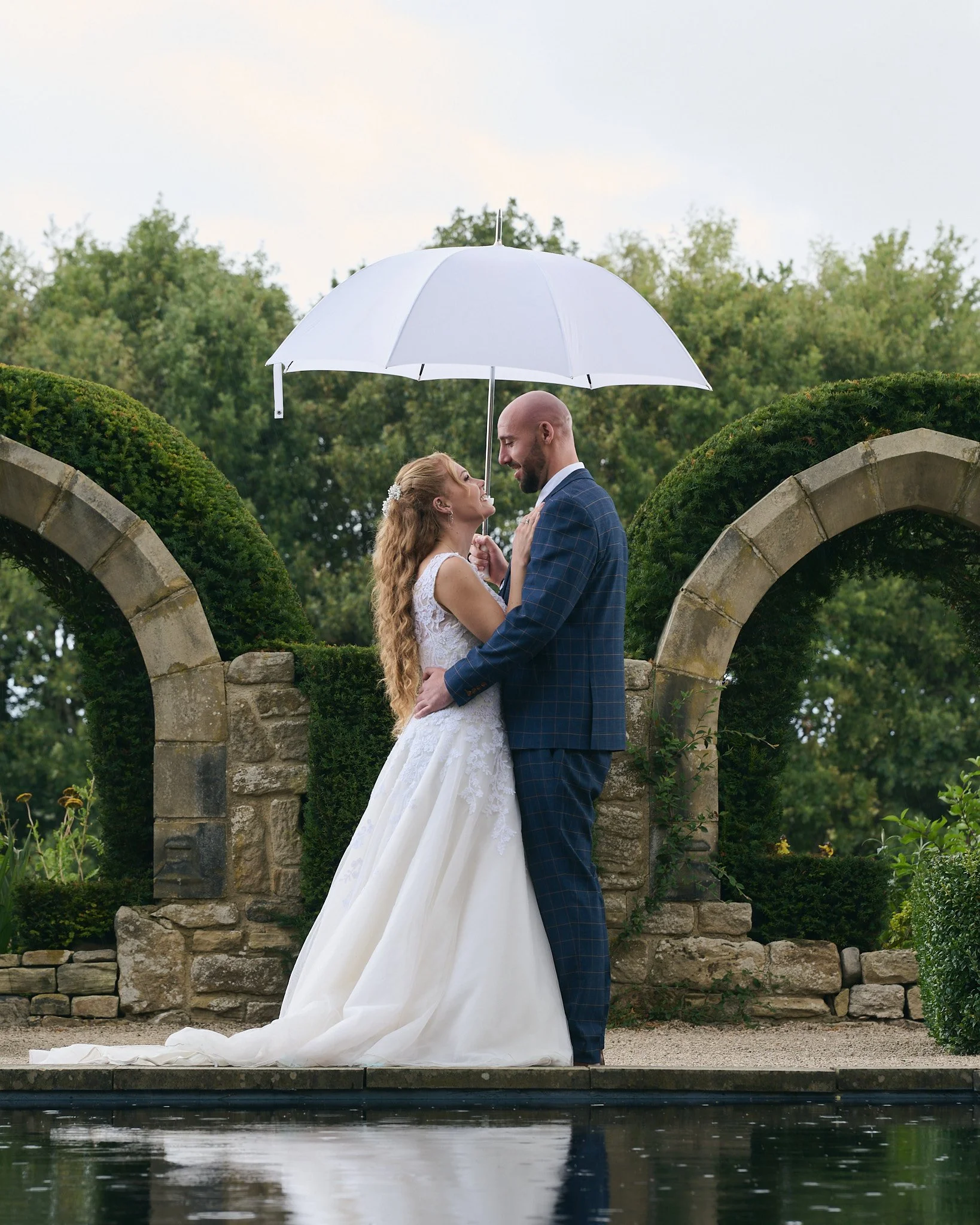 A bride and groom standing closely under a white umbrella, sharing a moment in an outdoor garden with stone arches and greenery.