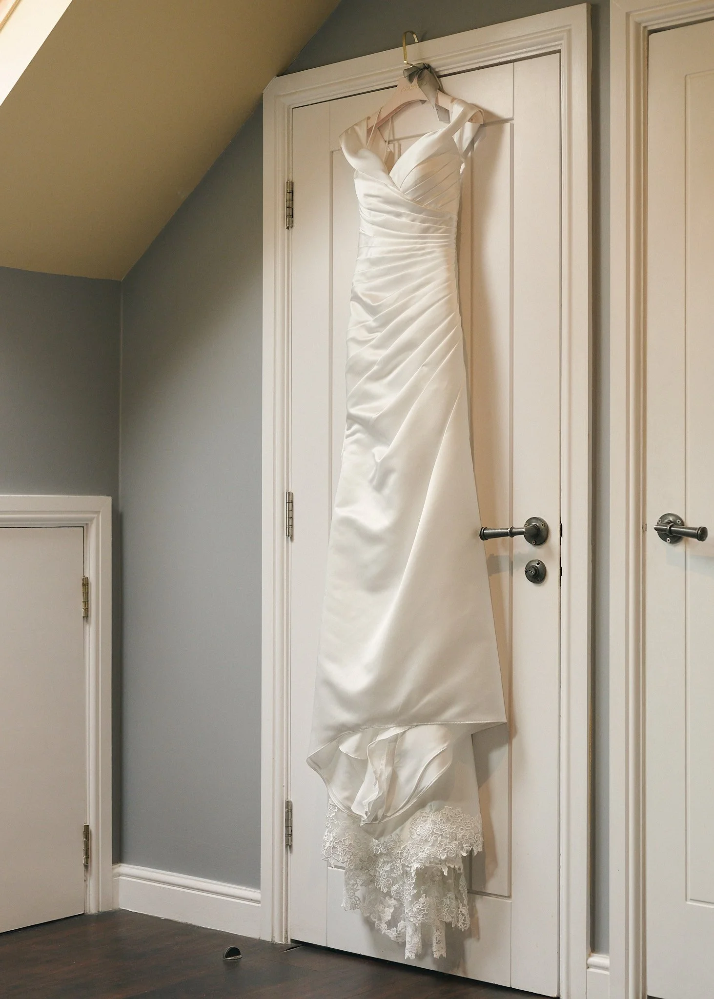 A white wedding dress hanging on a door hook in a room with gray and white walls and dark hardwood flooring.