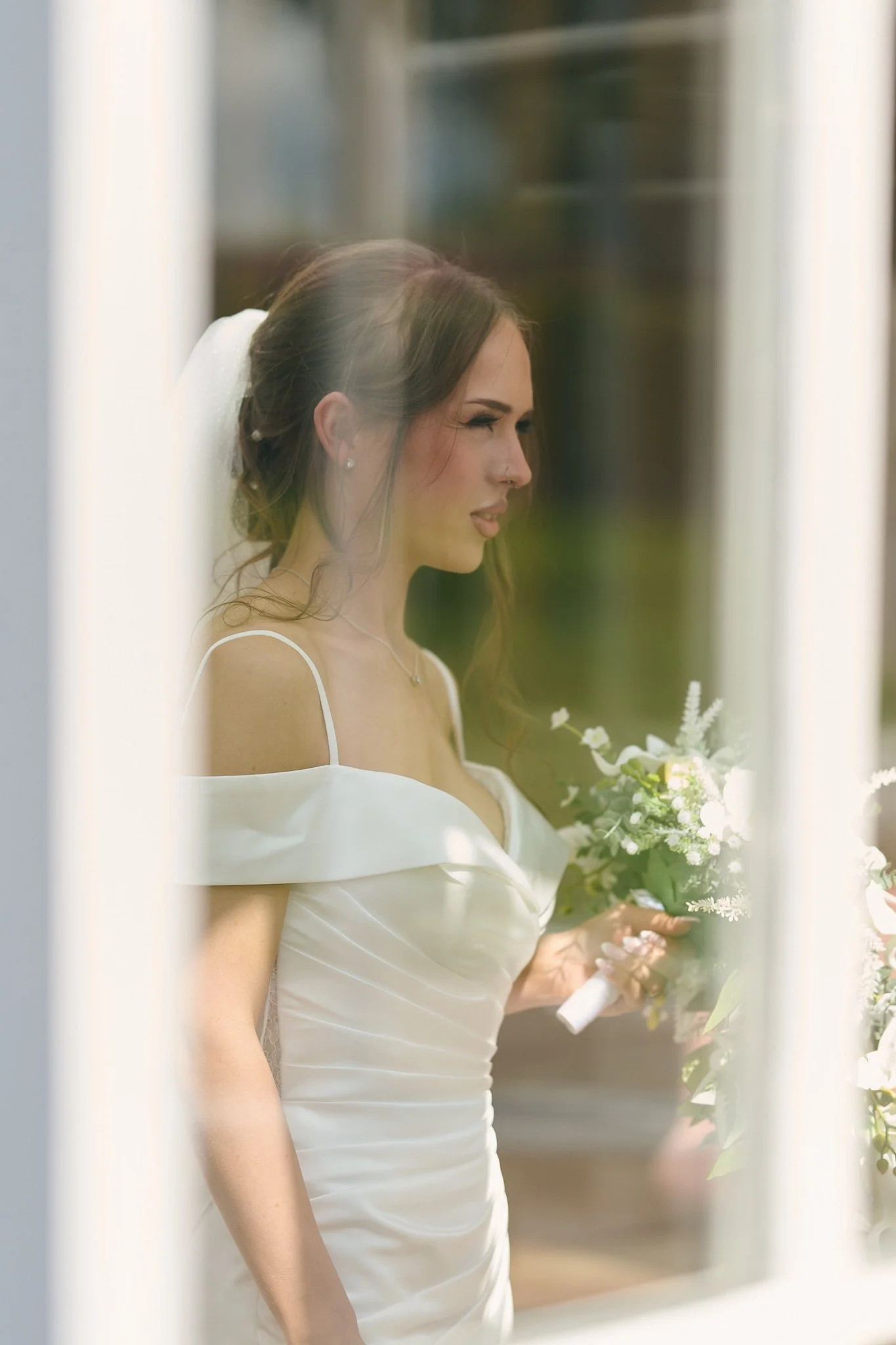 A bride with long brown hair styled in loose waves, wearing pearl earrings and a white off-shoulder wedding dress, holding a bouquet of white flowers and greenery, seen through a window or mirror with a blurred background.