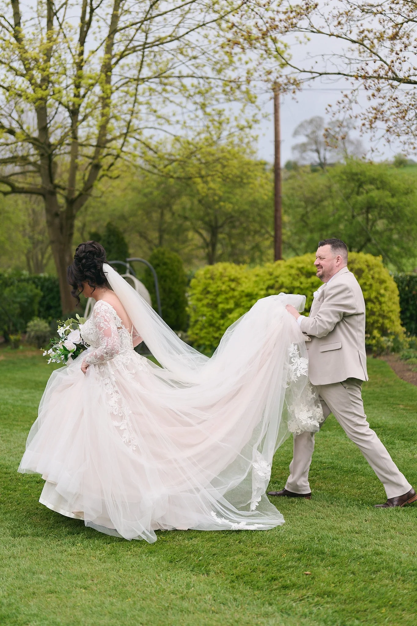 A bride and groom outdoors, with the groom holding the bride's train, surrounded by greenery and trees, on a cloudy day.