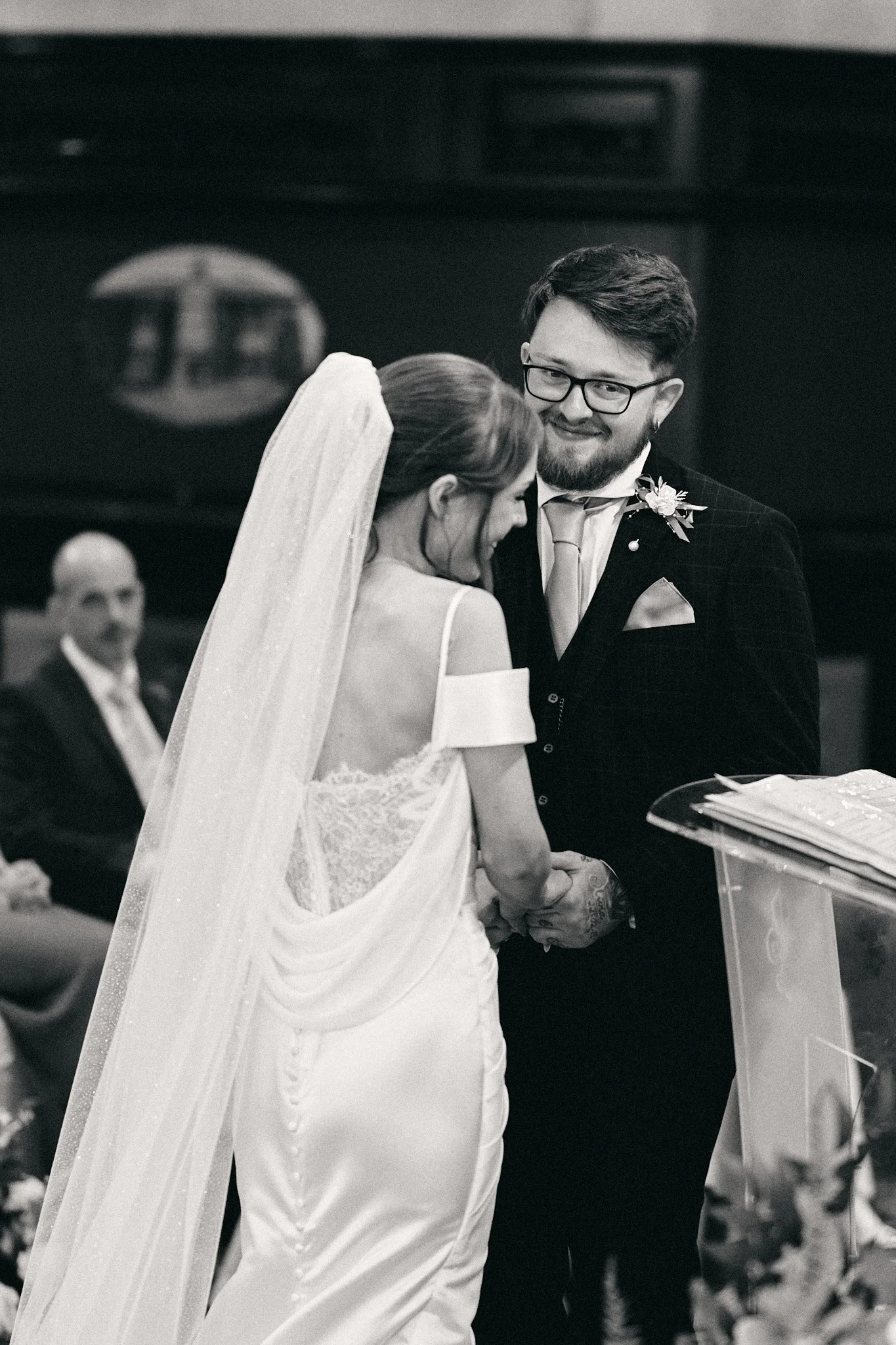 A black-and-white photo of a wedding ceremony showing a bride and groom holding hands, smiling, and looking at each other. The bride wears a white dress with lace details and a veil, while the groom wears a dark suit with a boutonnière and glasses.