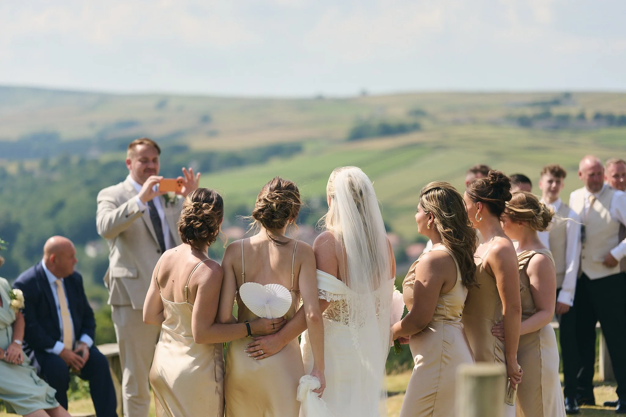 Group of brides and groomsmen at an outdoor wedding ceremony, with rolling green hills in the background.