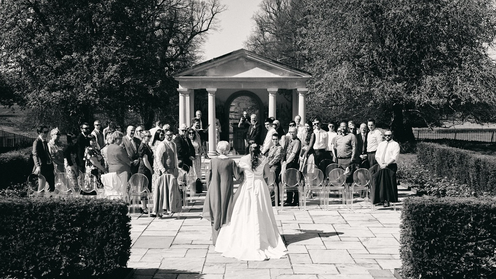 A wedding ceremony outdoors with a bride in a white gown standing before a group of guests, under a small pavilion with columns, surrounded by trees and garden.