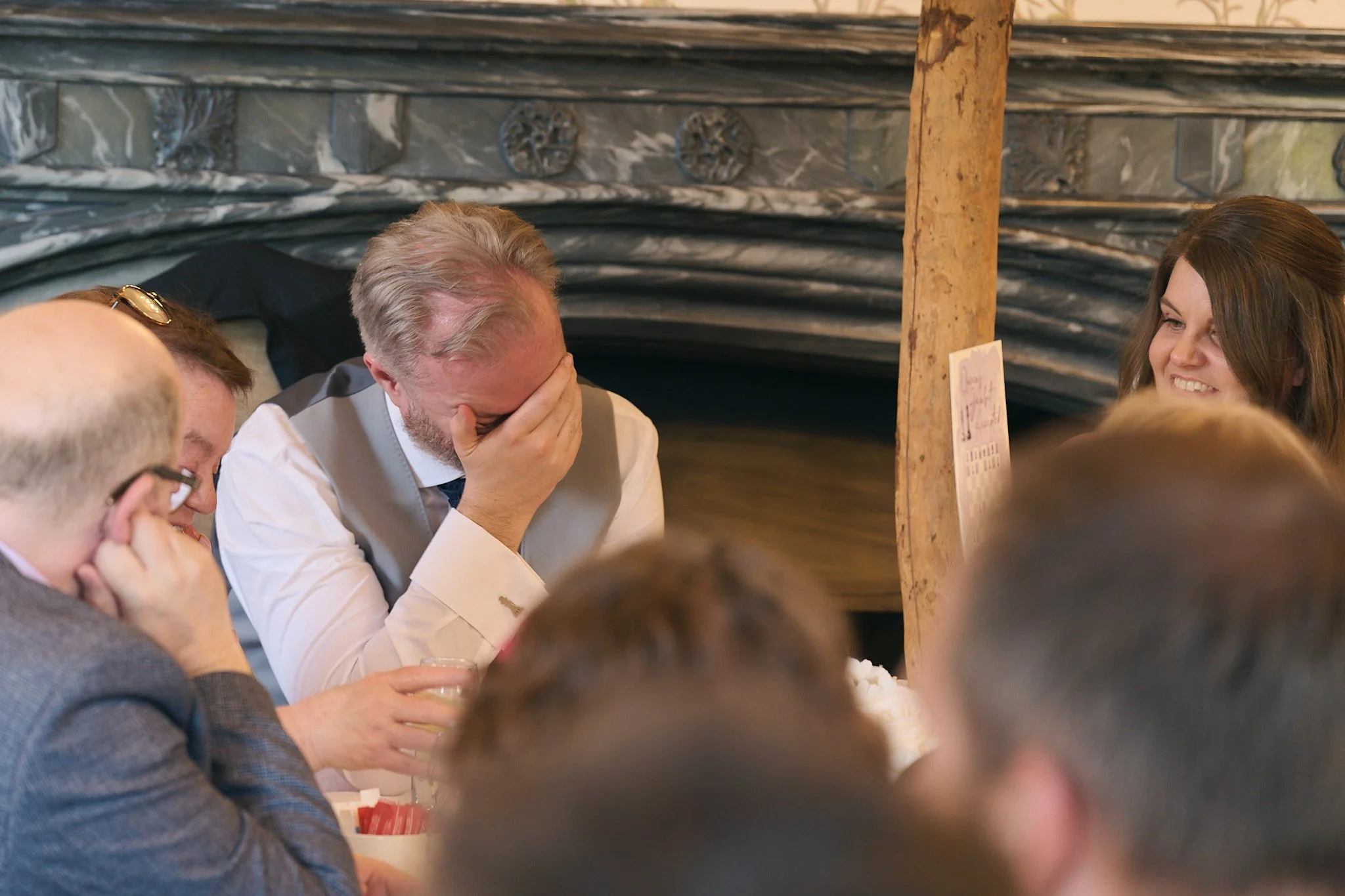 People sitting at a table, with one man covering his face and others smiling in an indoor setting.