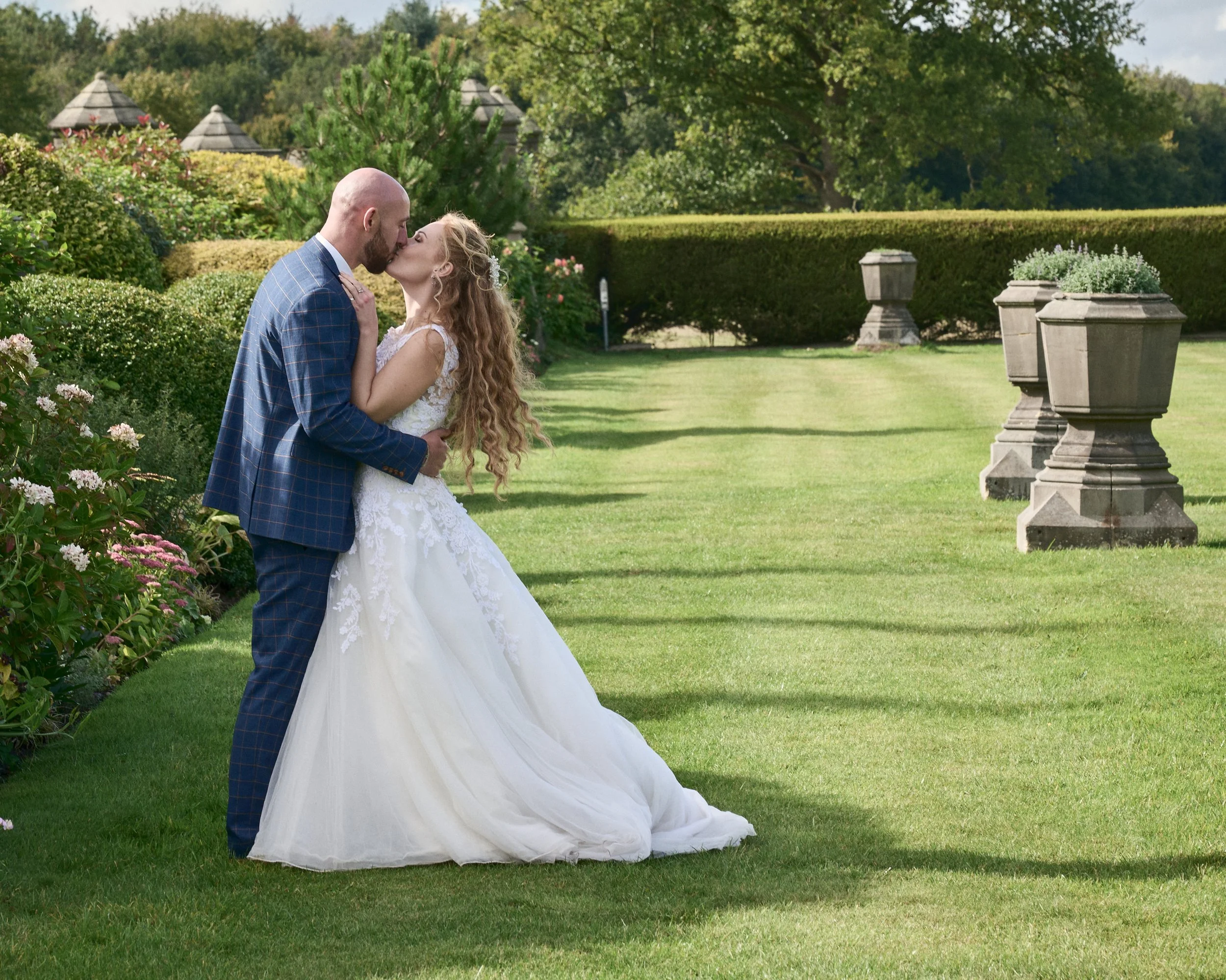 A bride and groom sharing a kiss outdoors on a sunny day, with greenery and decorative stone planters in the background.
