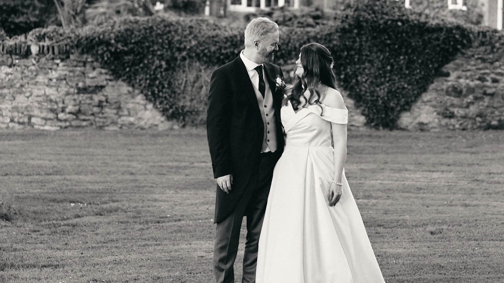 A man and woman dressed in formal attire, smiling and looking at each other, standing outdoors on a lawn with a stone wall and bushes in the background.
