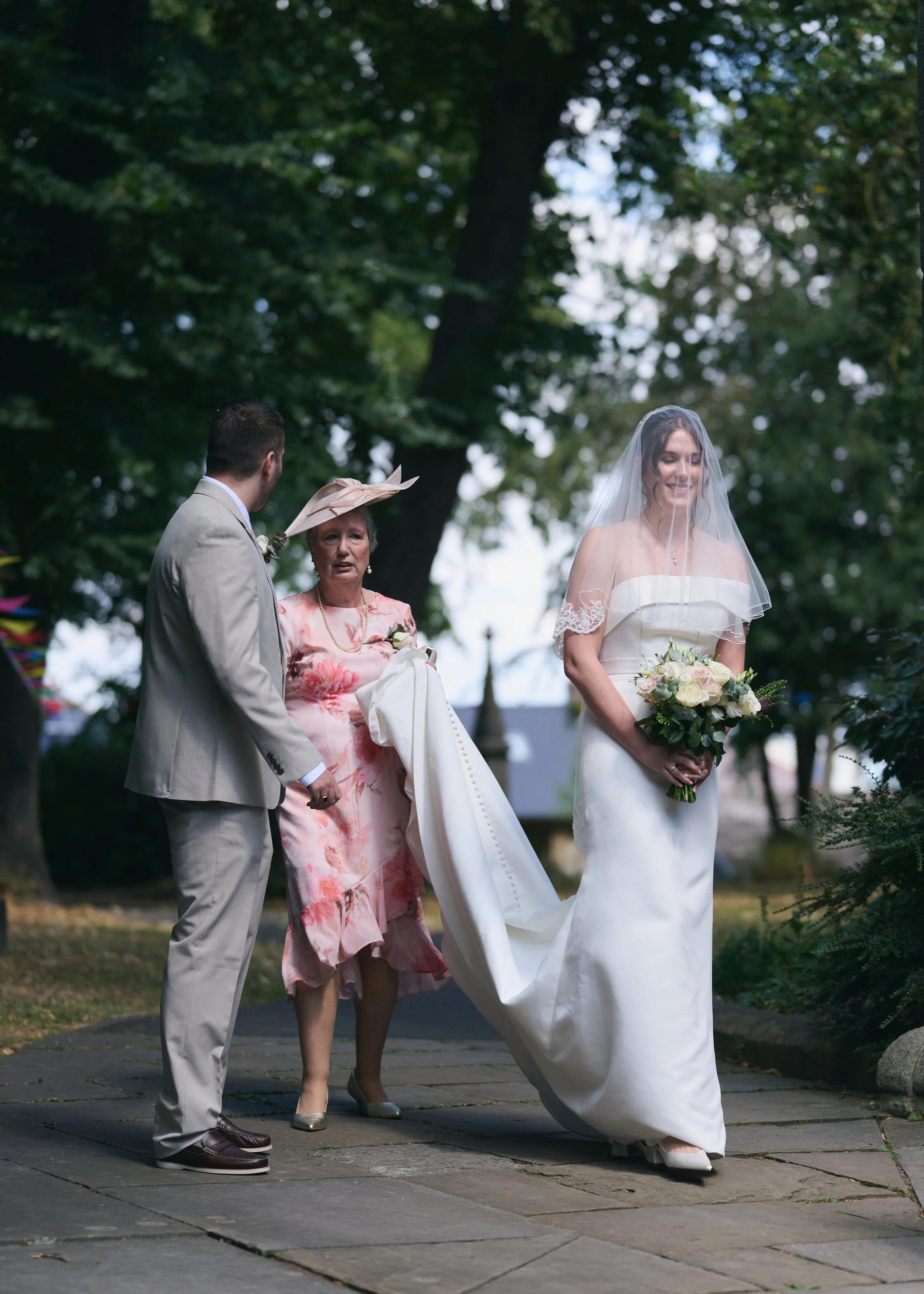 A bride in a white wedding dress with a veil holding a bouquet of flowers stands outside on a pathway, smiling. An older woman in a pink floral dress and hat, and a man in a light suit, are with her, possibly during a wedding ceremony in a park or garden setting.