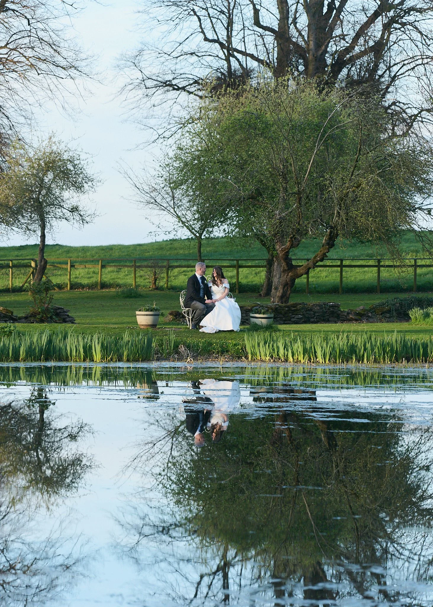 A couple in wedding attire sitting on a white bench beside a pond, with trees in the background and their reflection visible in the water.