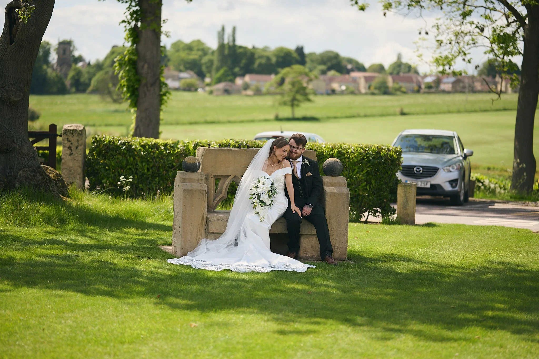 A newlywed couple sitting on a stone bench in a park, surrounded by green grass and trees. The bride is wearing a white wedding dress and veil, and holding a bouquet of white flowers. The groom is dressed in a black suit with a white shirt and tie, smiling. Two cars are parked behind them, and there are trees and a distant village or town in the background.