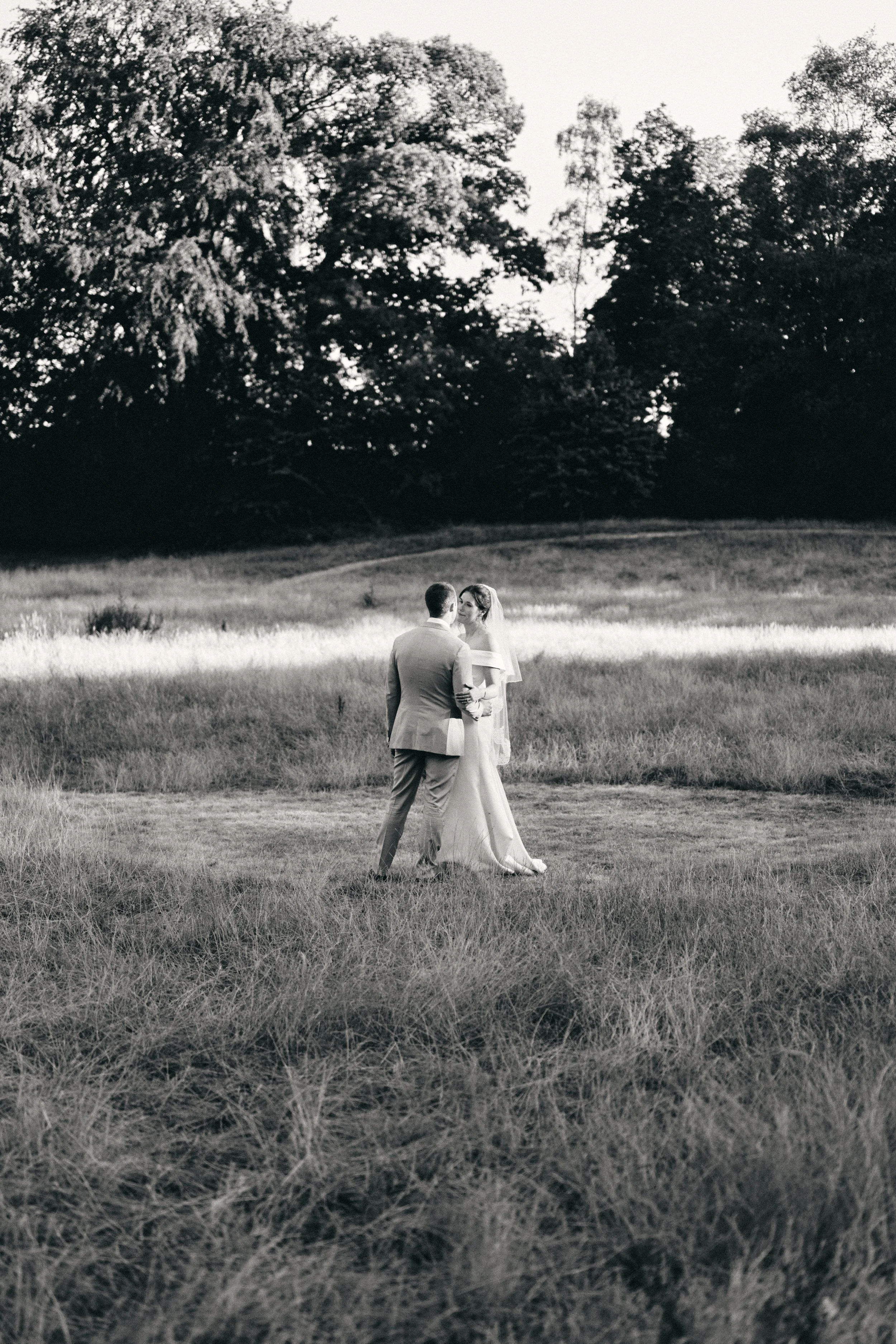A black and white photo of a bride and groom standing in a grassy field, with trees in the background.
