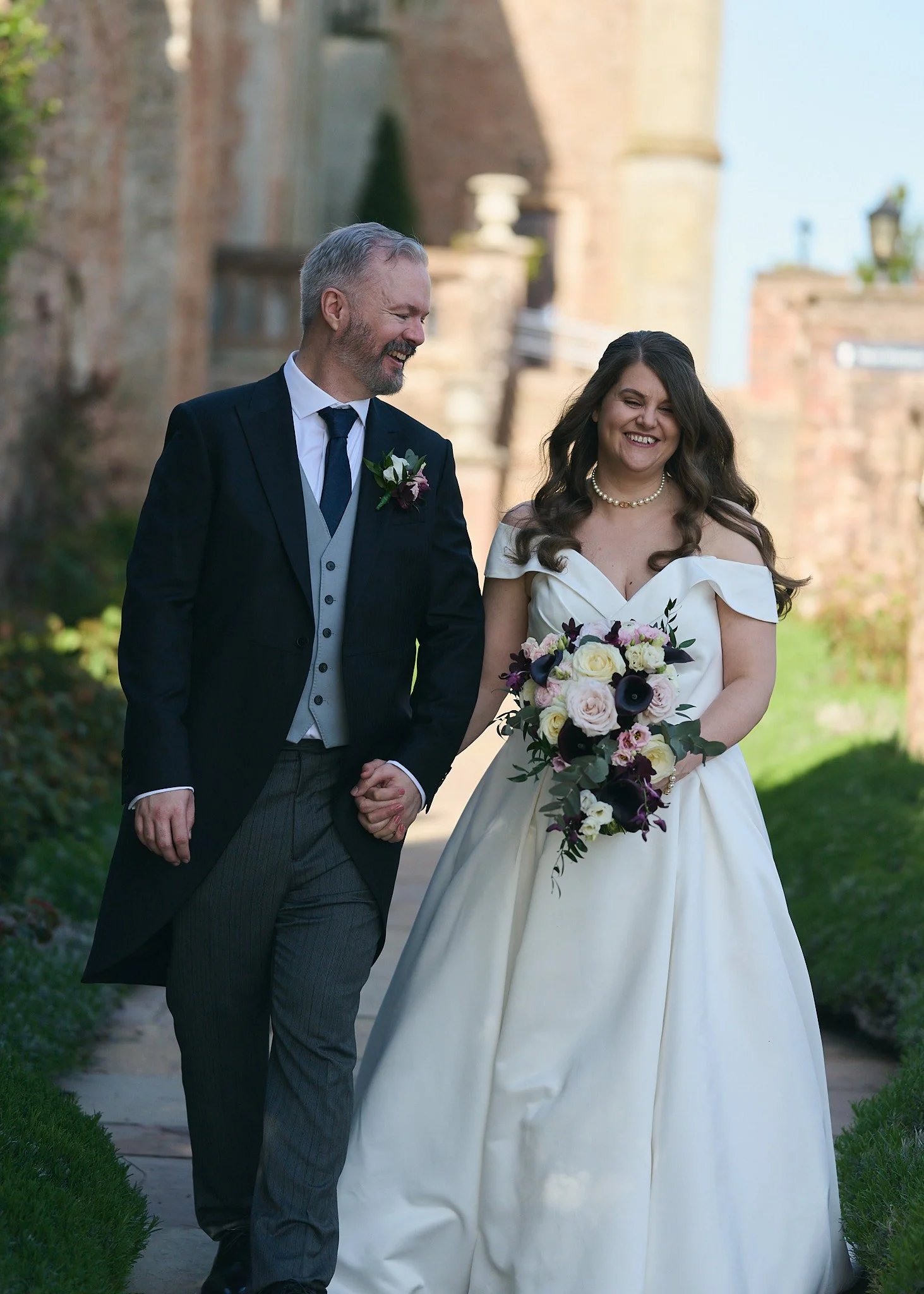 A happy couple, dressed in wedding attire, walking outdoors holding hands, with the man in a dark suit and the woman in a white wedding gown holding a bouquet of flowers, smiling and enjoying each other's company.