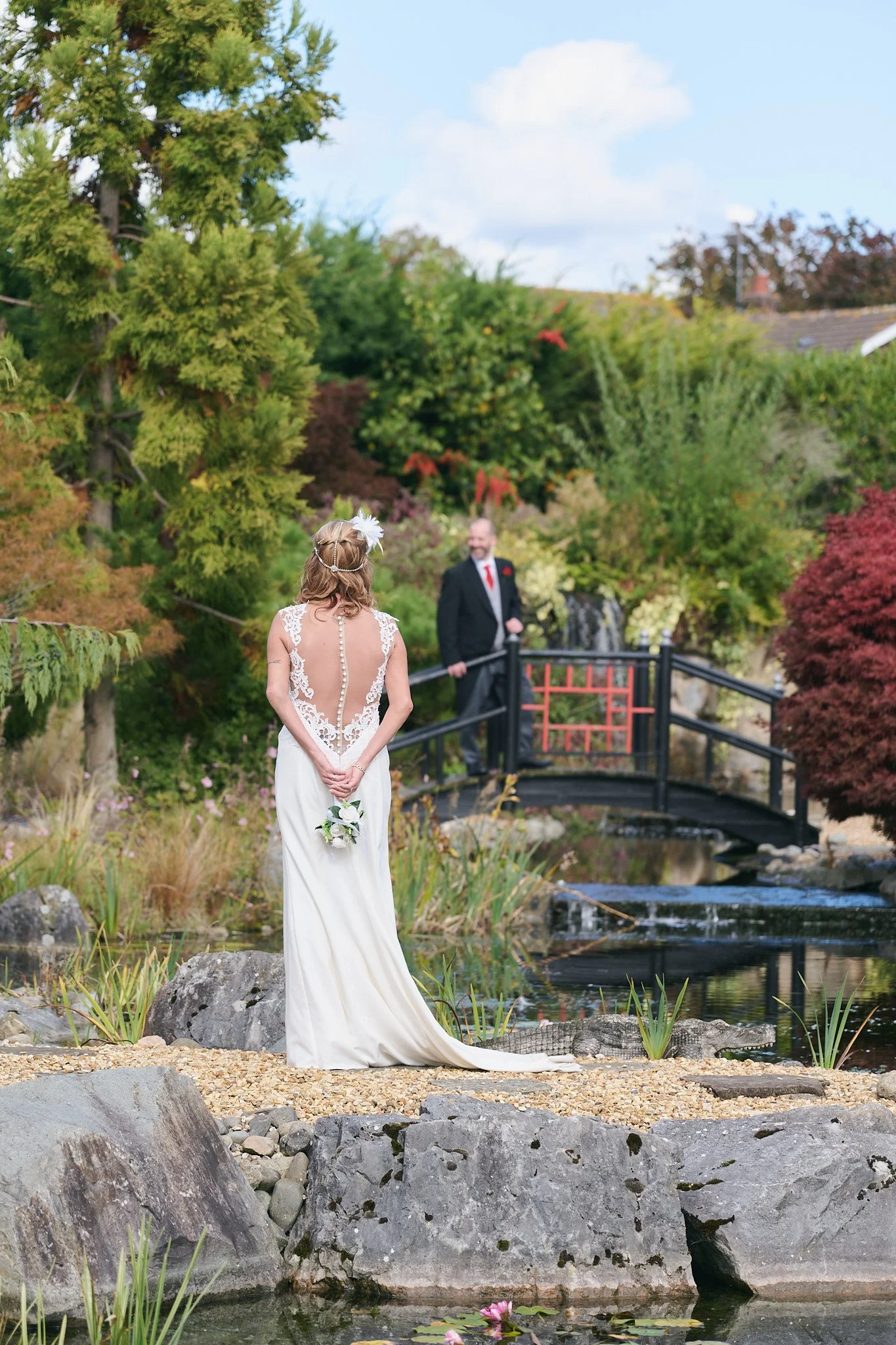 Bride in a white wedding dress with lace back facing a man in a suit by a small decorative pond with pink water lilies, surrounded by rocks and lush green trees, on a bright day.