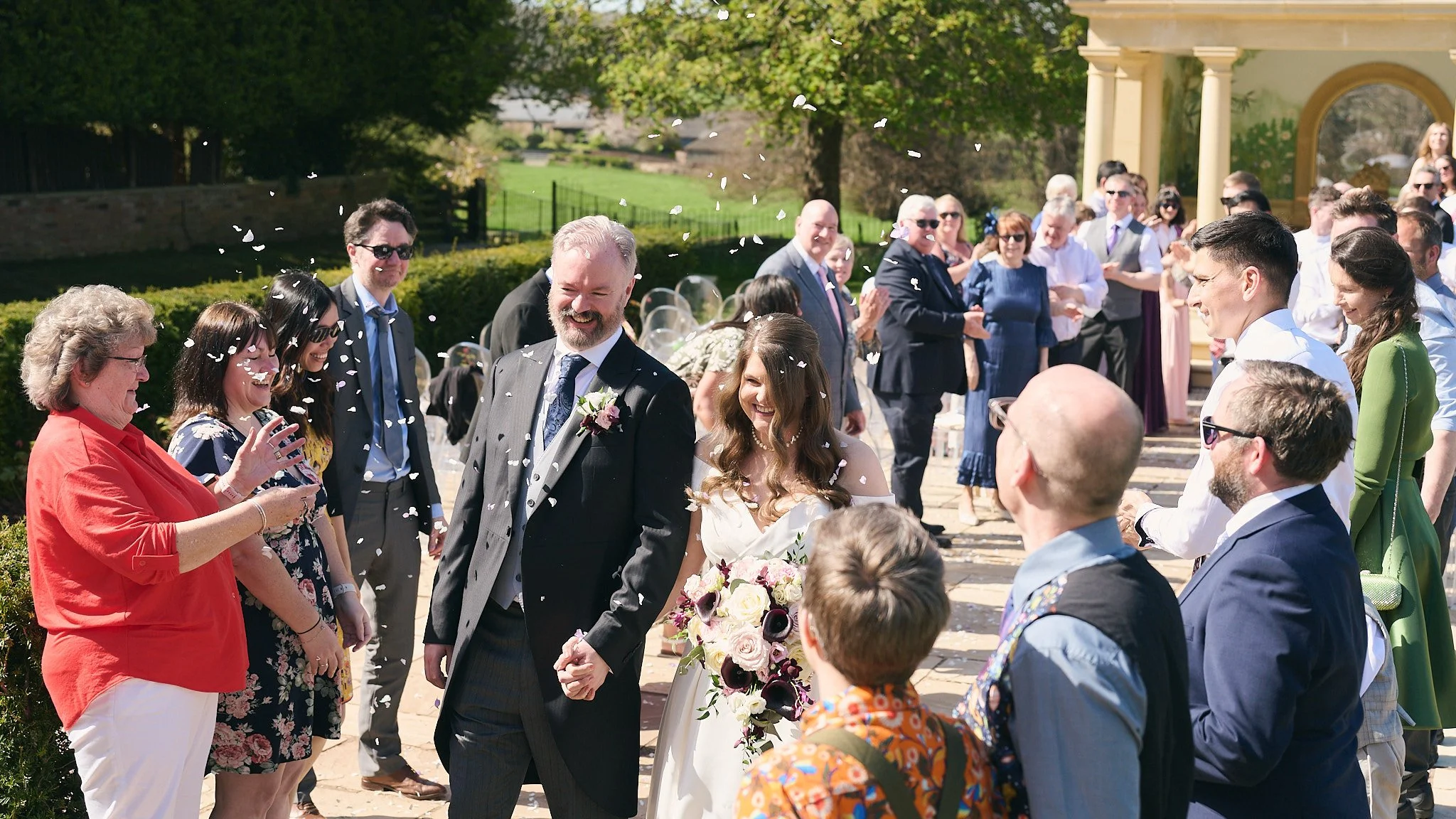 A wedding celebration outdoors with the bride and groom walking hand in hand, surrounded by guests throwing white confetti on a sunny day.