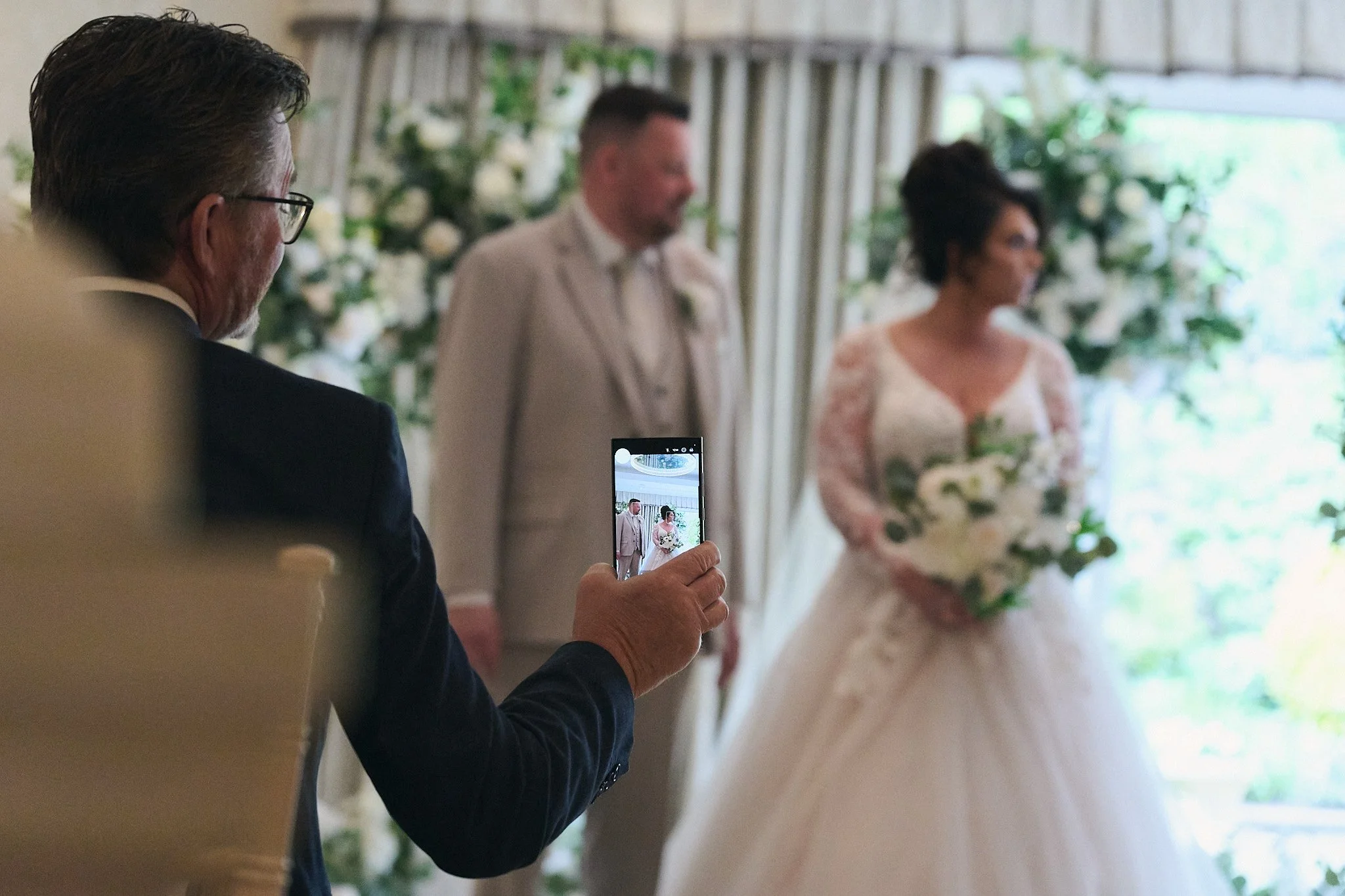A man in a black suit with glasses is taking a photo of a bride and groom during a wedding ceremony. The bride is wearing a lace wedding dress and holding a bouquet, and the groom is dressed in a light-colored suit. They are standing in front of a floral backdrop.