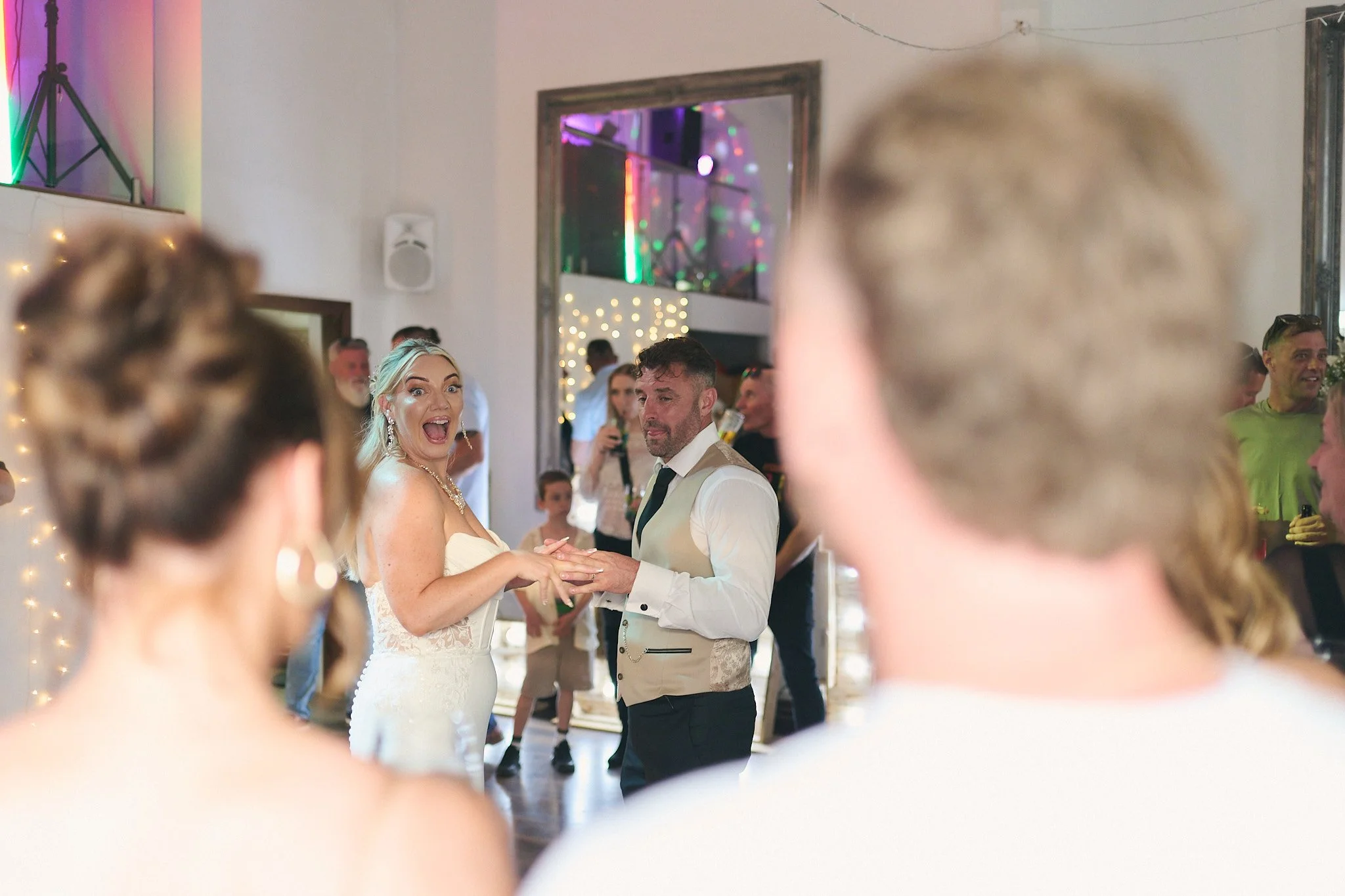 A bride and groom dancing at their wedding reception, surrounded by guests and festive decorations with colorful lighting.