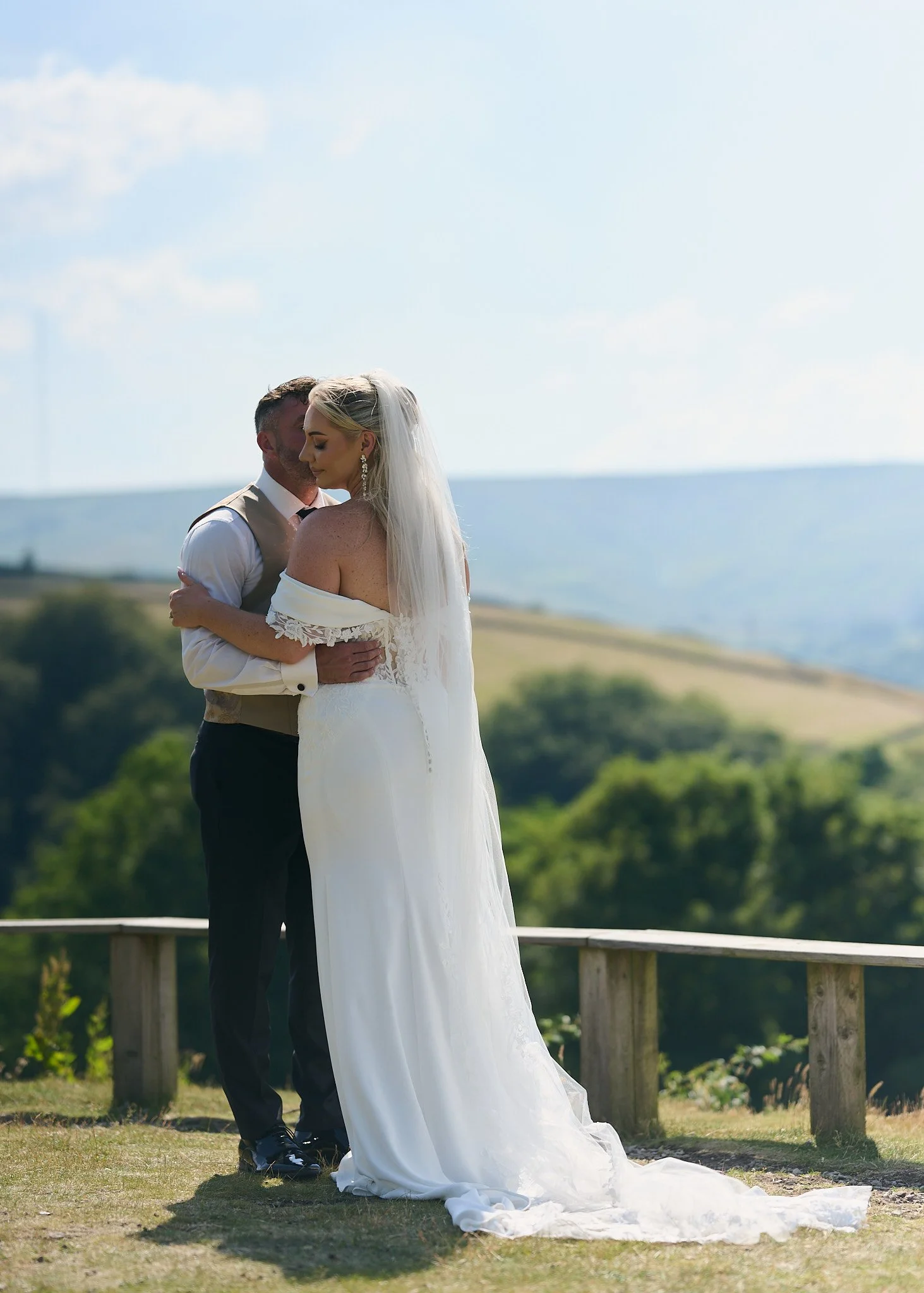 A bride and groom embracing outdoors on a sunny day, with the ocean visible in the background.