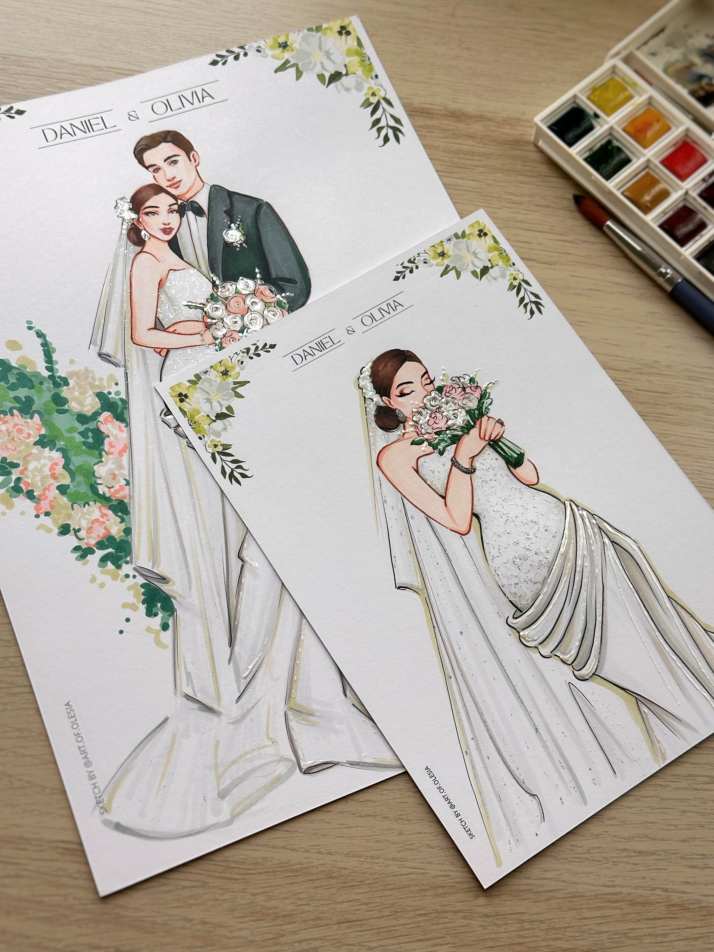 Illustration of a bride and groom on wedding stationery with floral designs, watercolor style, on a wooden table, near a watercolor paint set and brush.