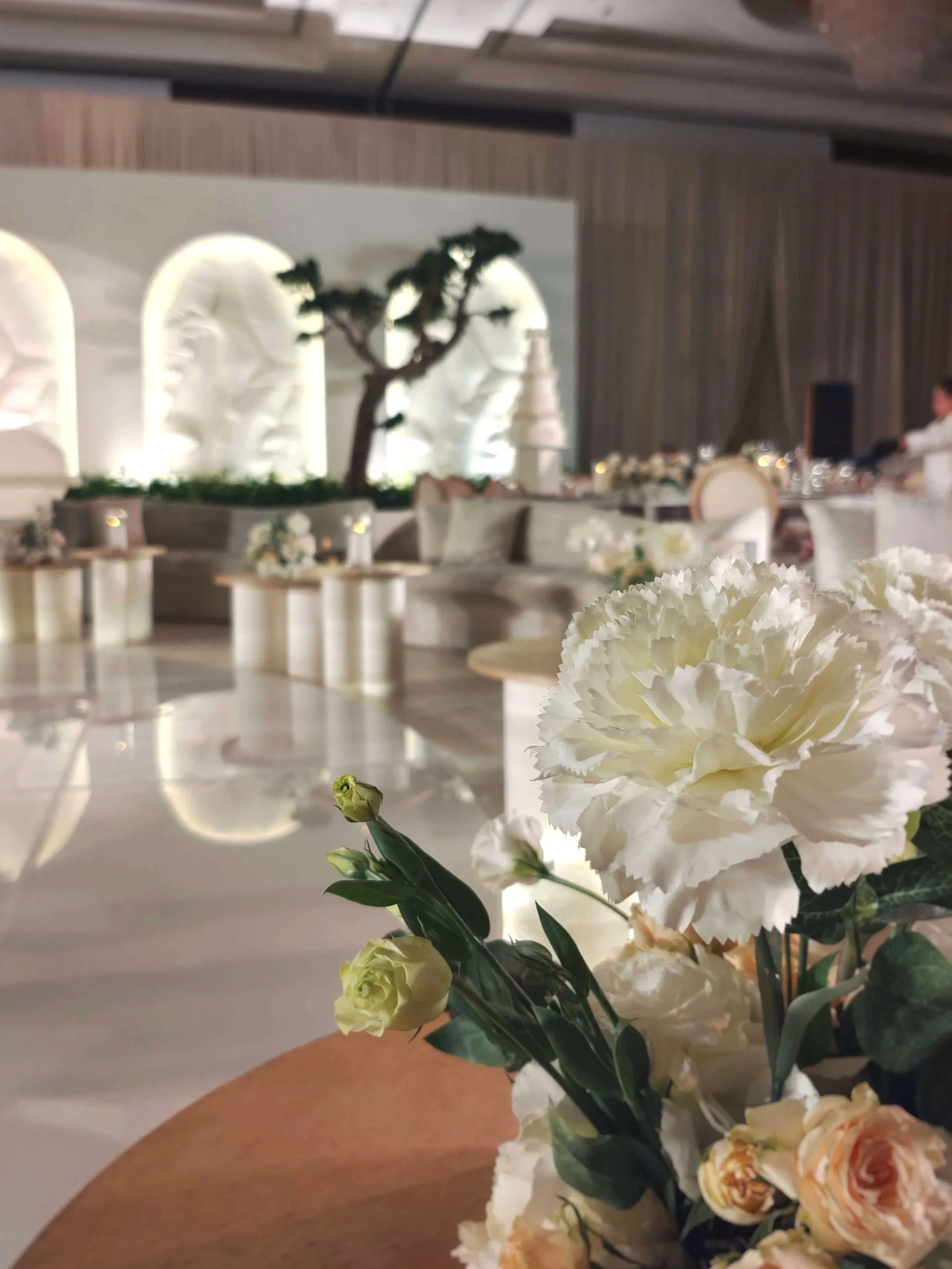 A bouquet of white and peach-colored flowers, including carnations and roses, held in front of a banquet hall decorated with a tree sculpture and elegant tables.