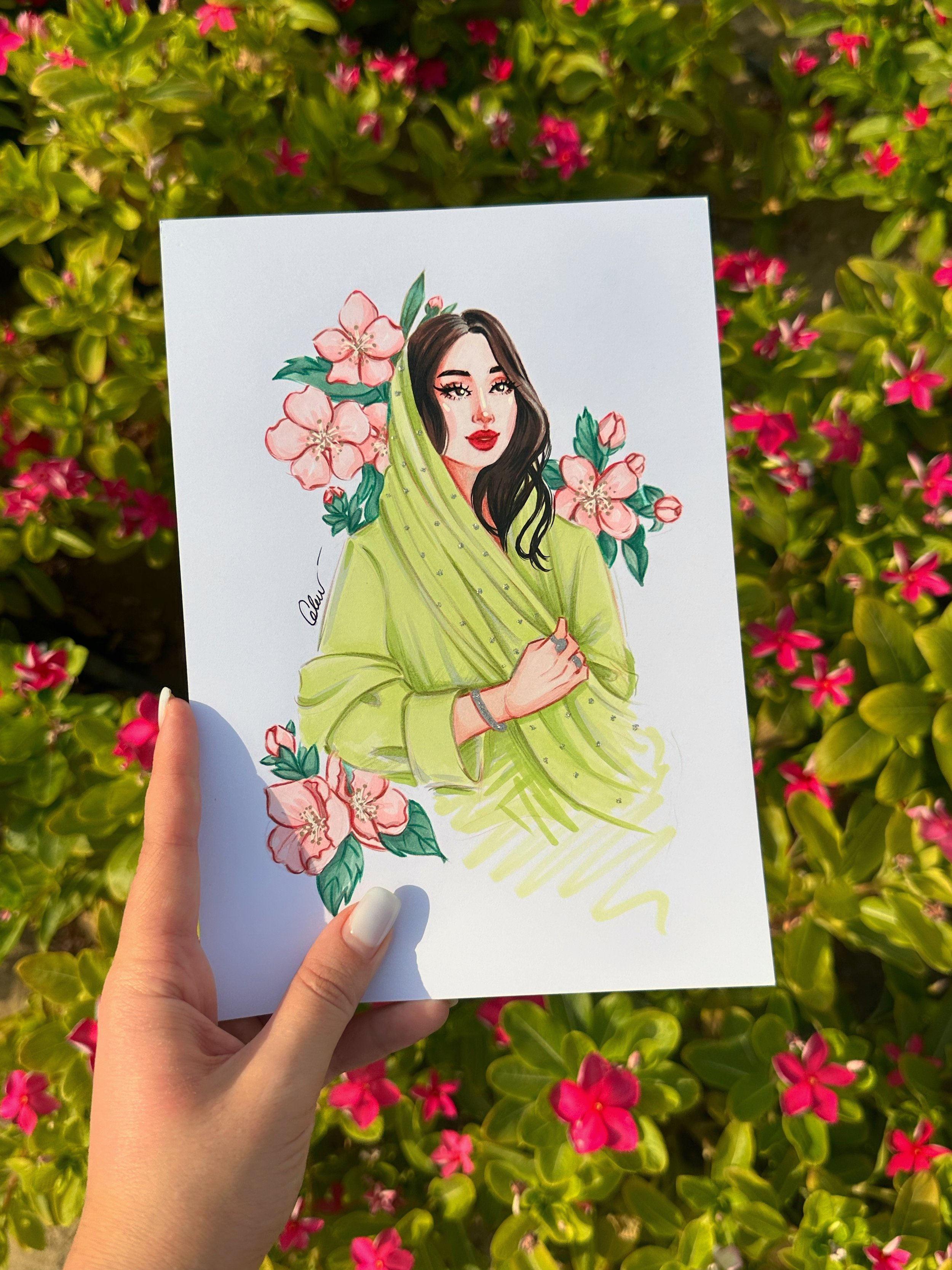 A hand holding a drawing of a woman with dark hair, red lips, and green attire, surrounded by pink flowers and green leaves, with pink flowers in the background.