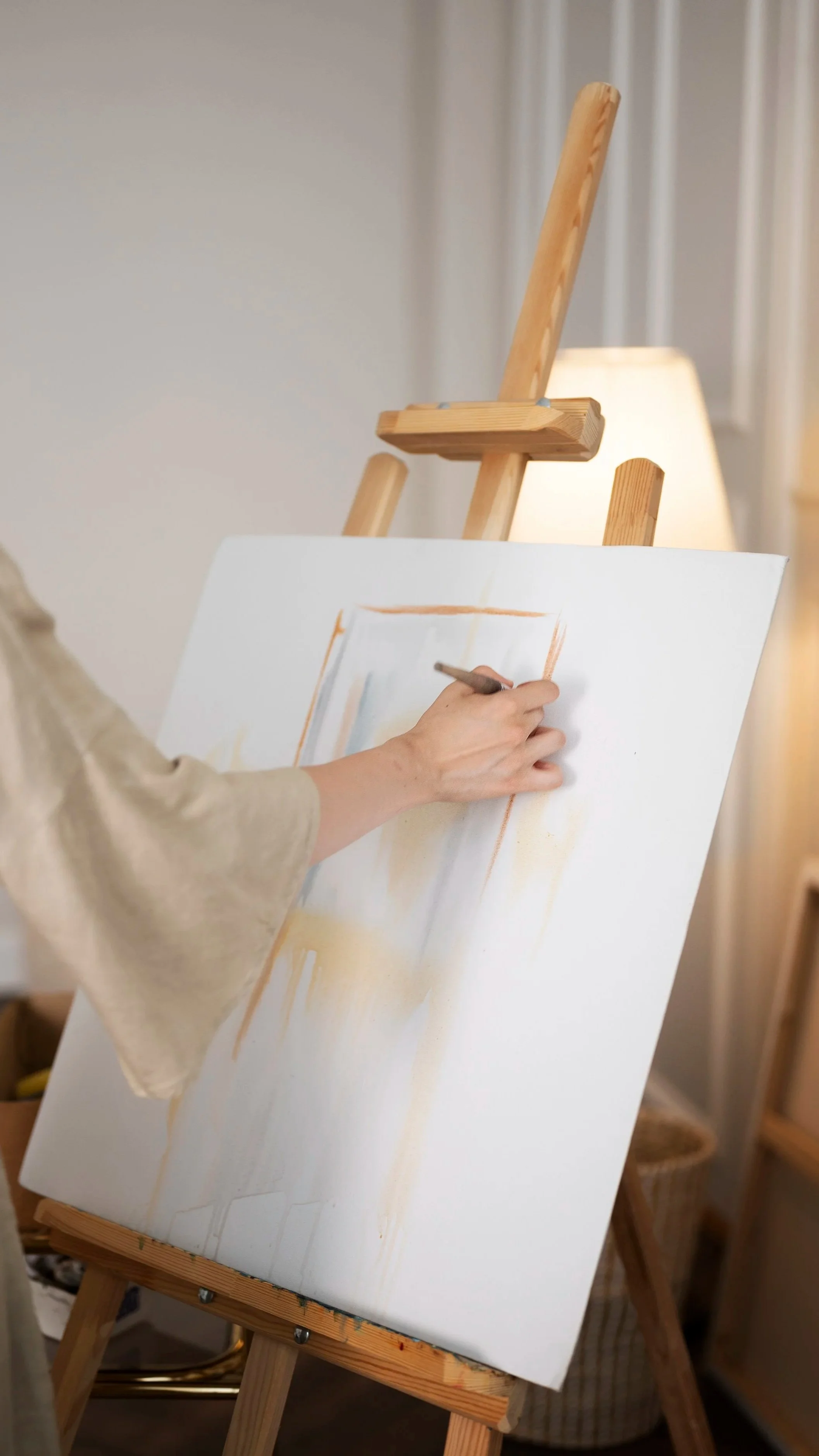 Person painting a canvas on an easel with a brush, indoors with soft lighting.
