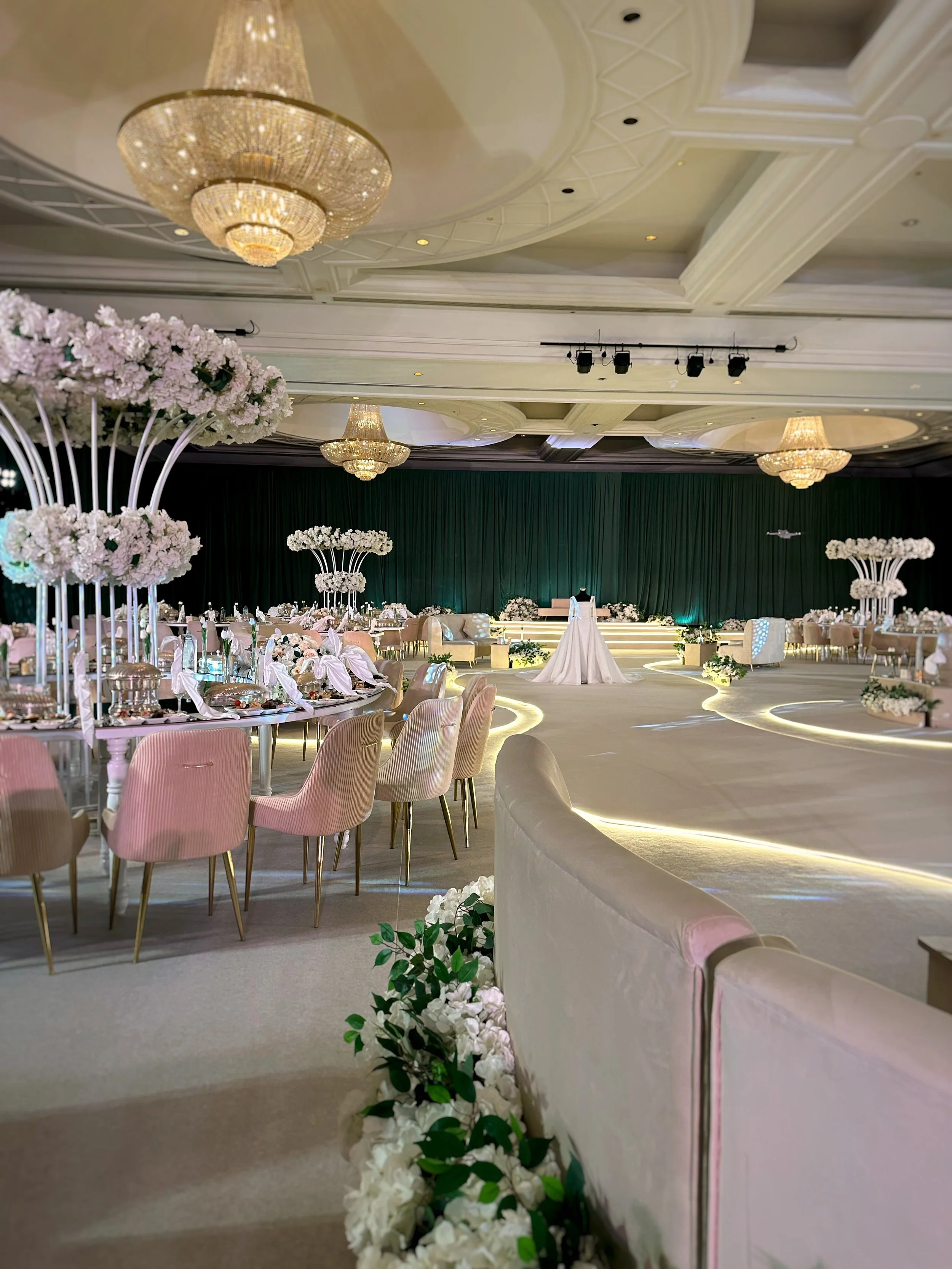An elegant banquet hall decorated for a wedding reception with pastel pink chairs, tall floral centerpieces, chandeliers, and a stage with a green curtain backdrop.
