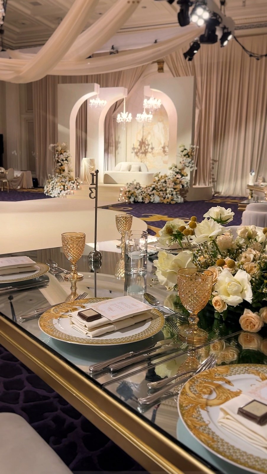 Elegant wedding reception table setup with gold-rimmed plates, floral centerpieces, and a stage decorated with flowers and drapery in the background.