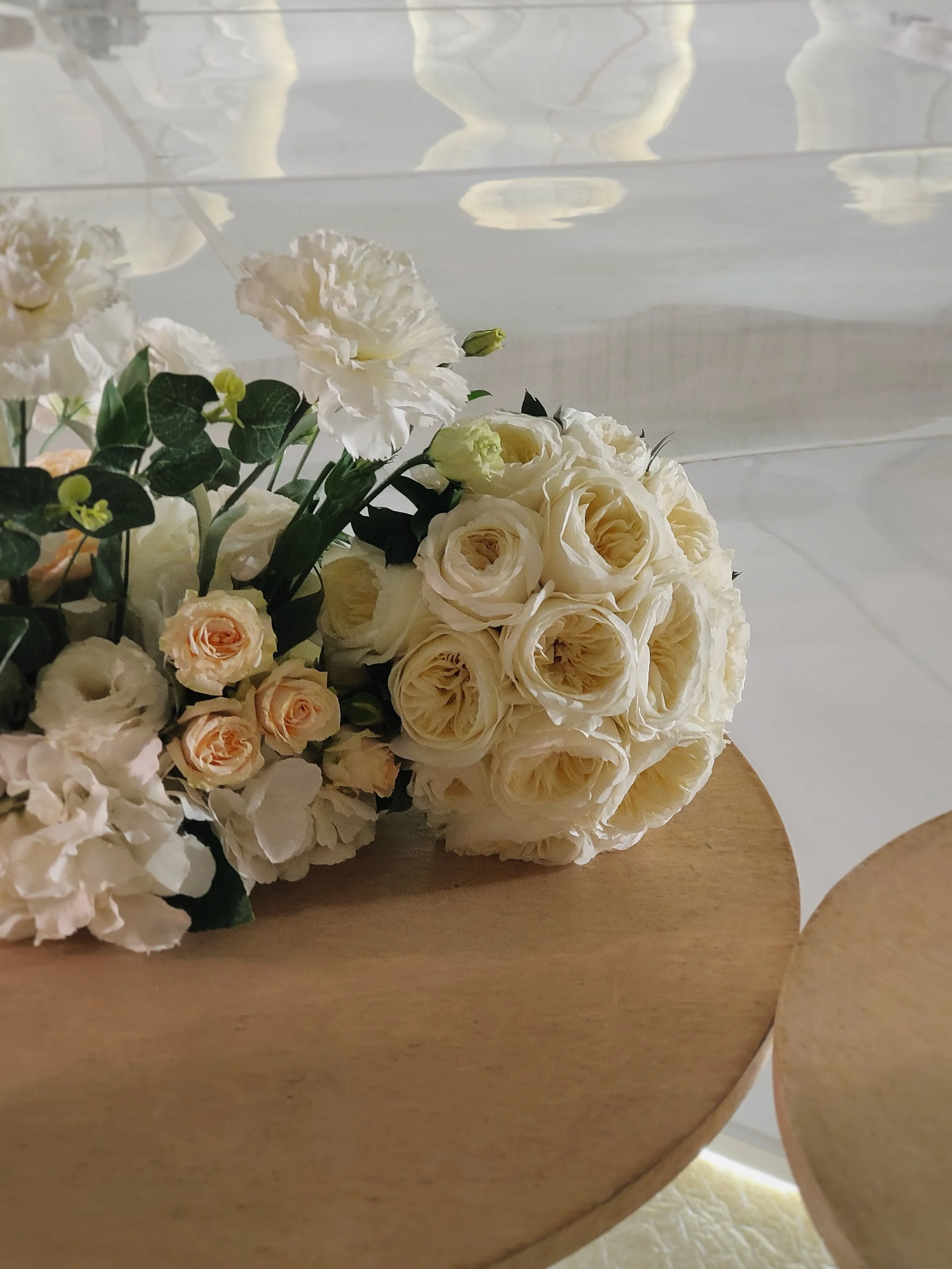 A wedding bouquet of cream-colored and pale peach roses with white carnations and greenery placed on a wooden table.